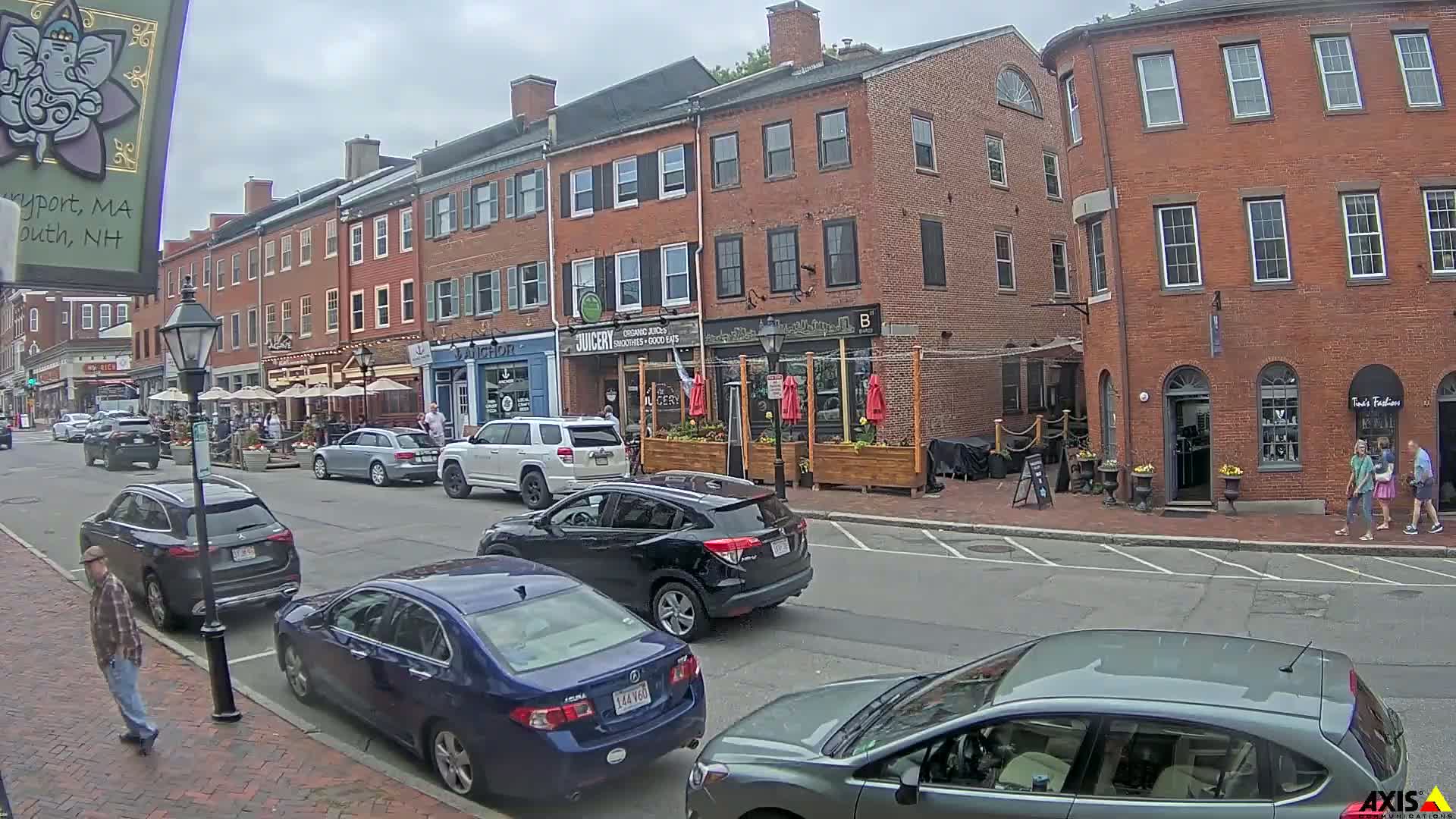 A street scene on an overcast day shows several parked cars and pedestrians in front of brick buildings with outdoor seating areas.