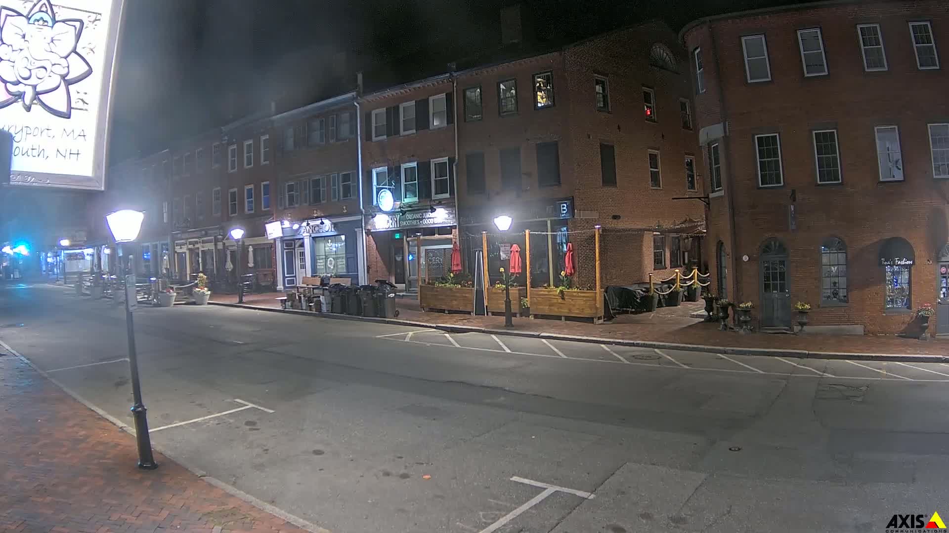 A nighttime street scene shows several brick buildings with outdoor seating areas, and streetlights illuminating an empty road.