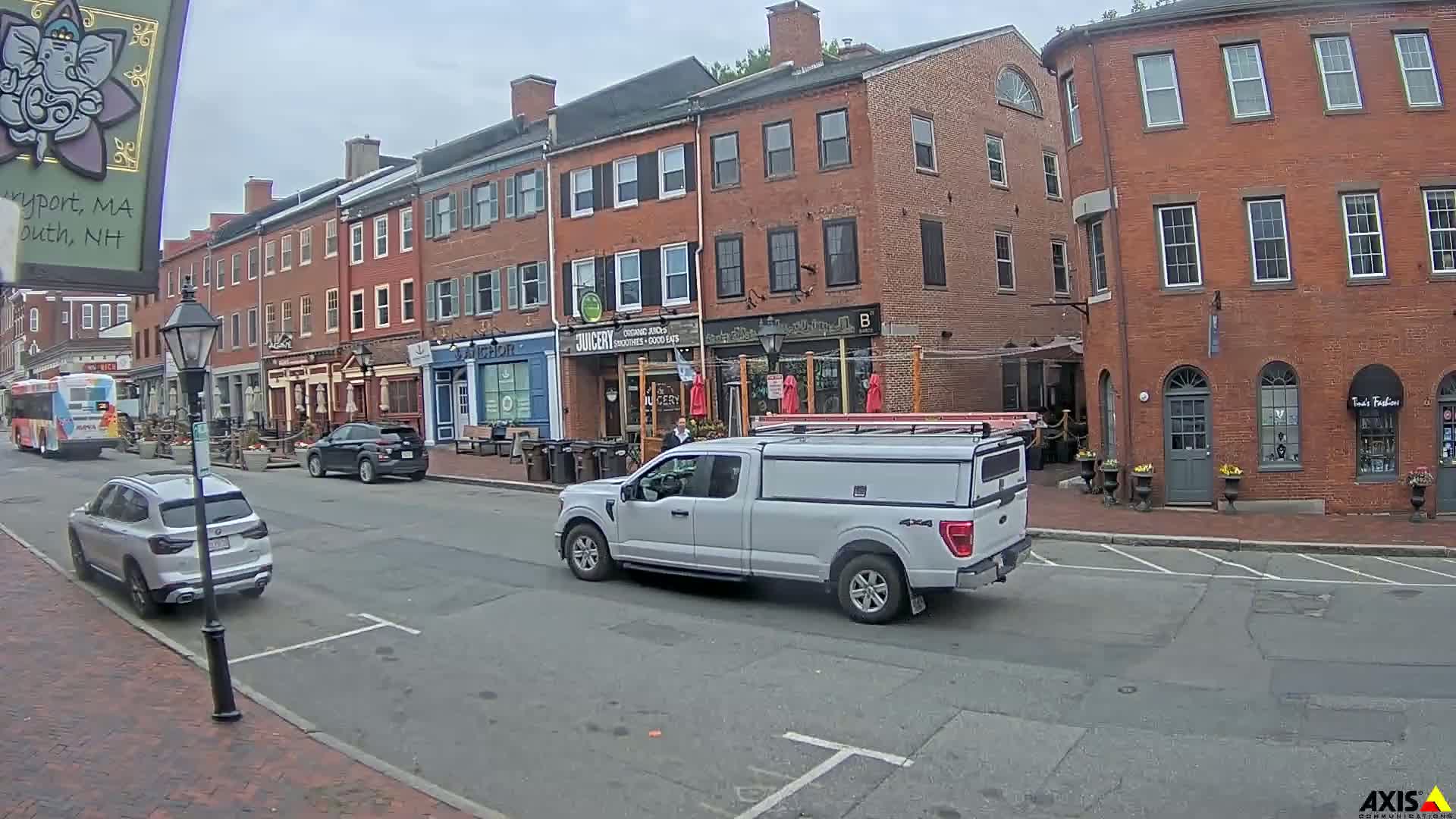 A light-grey pickup truck drives down a street lined with brick buildings on an overcast day.