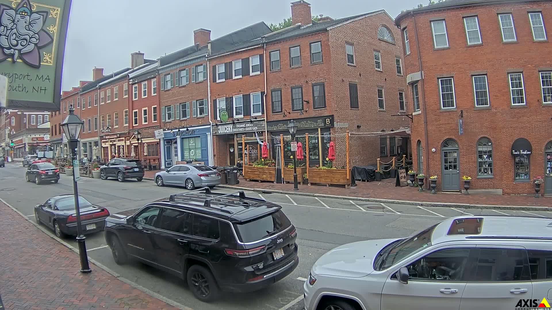 A street scene on an overcast day shows several parked cars in front of brick buildings with various storefronts, including a juice bar and a clothing store.
