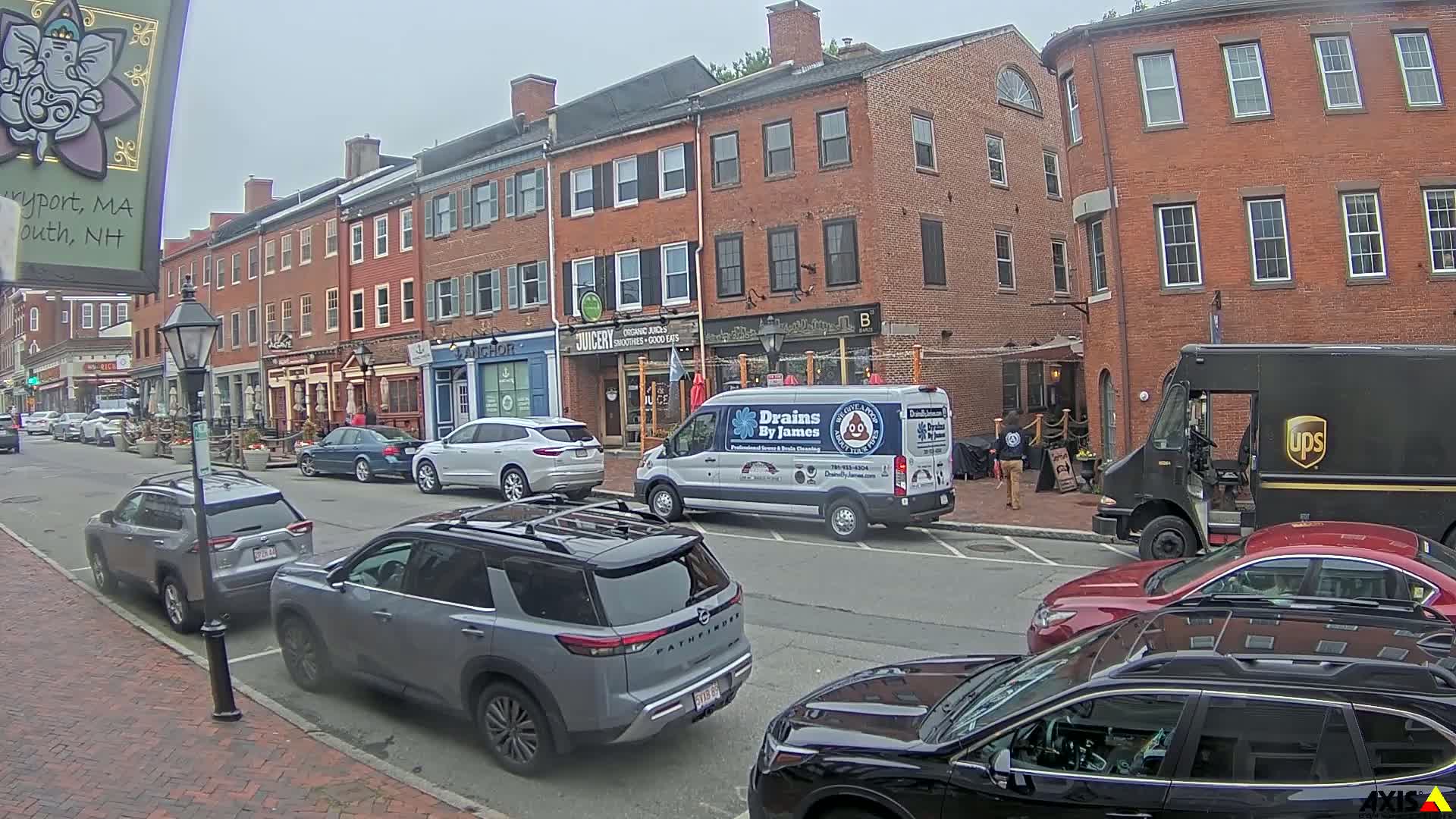 A street scene on an overcast day shows several parked cars in front of brick buildings with businesses on the ground floor.