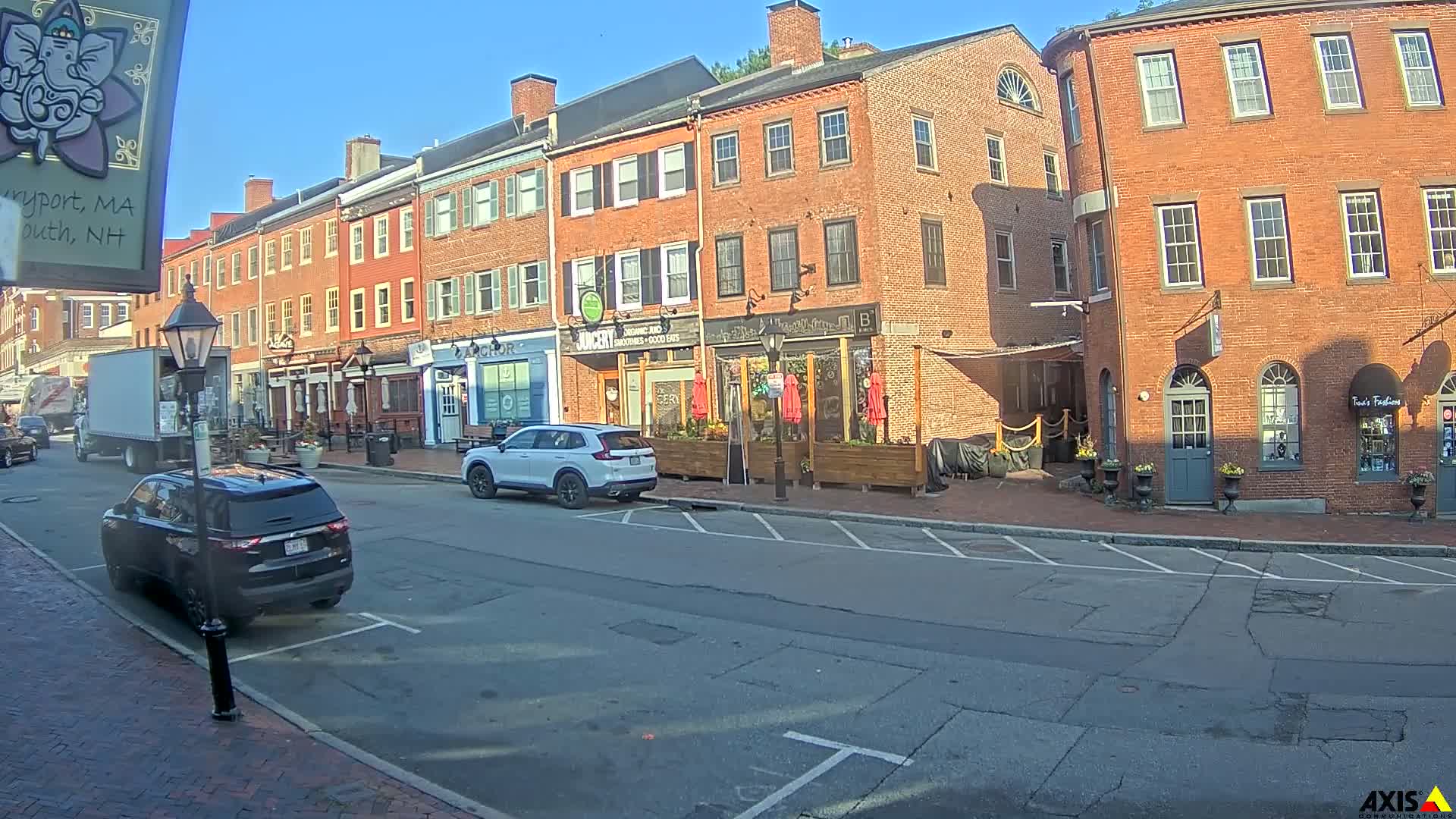 A sunny day on a brick-lined street shows several parked cars, a delivery truck, and multiple storefronts with outdoor seating areas.