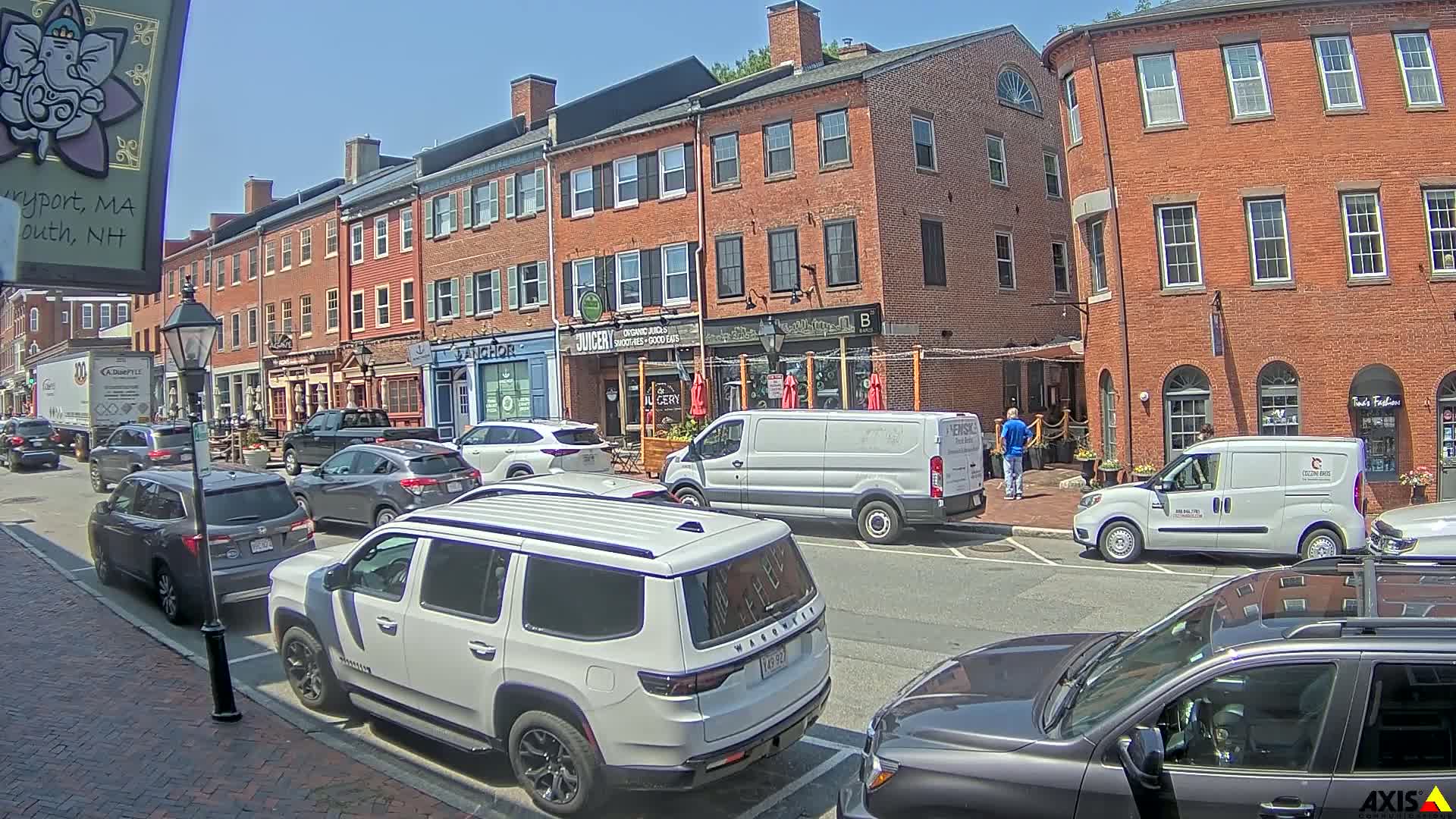A sunny day on a brick-lined street with parked cars and several people near shops and restaurants.