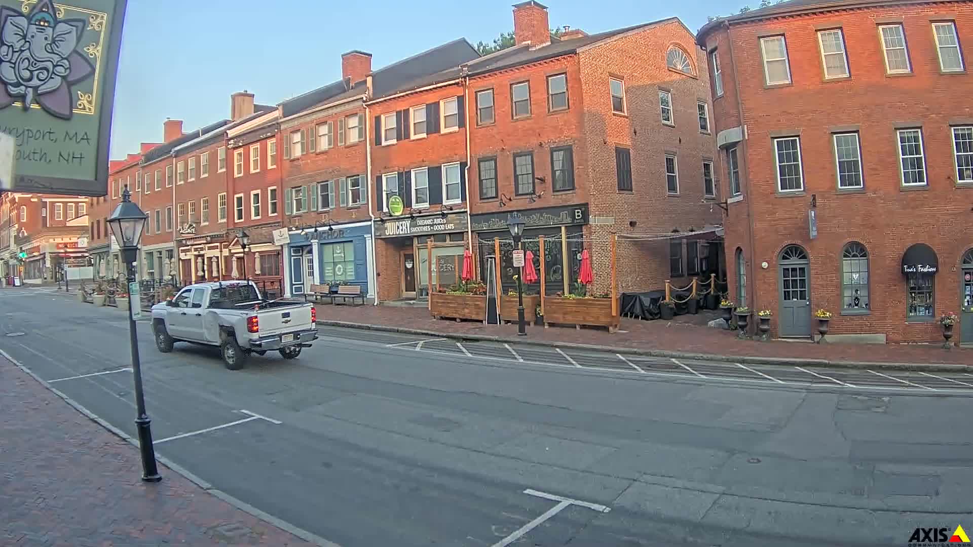A light-colored pickup truck drives down a street lined with brick buildings on a clear, sunny day.