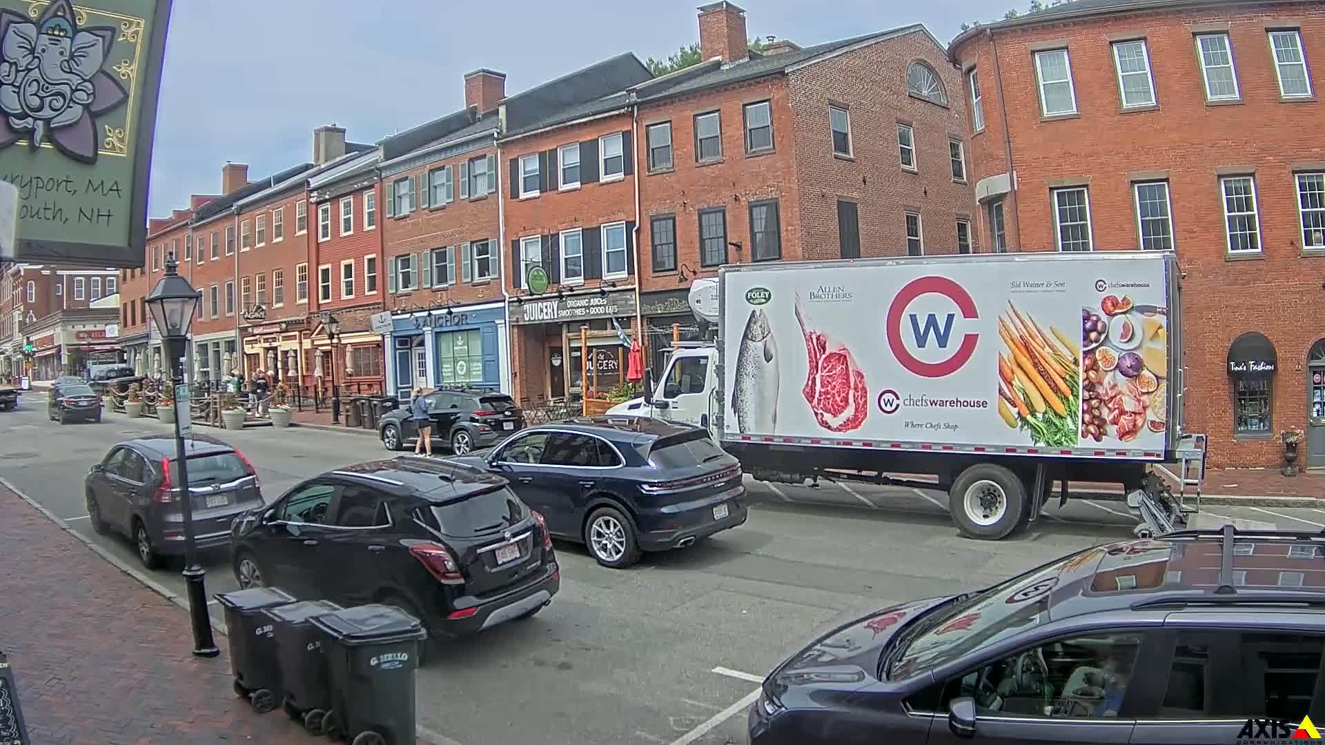 A city street with parked cars and a food delivery truck, under a partly cloudy sky.