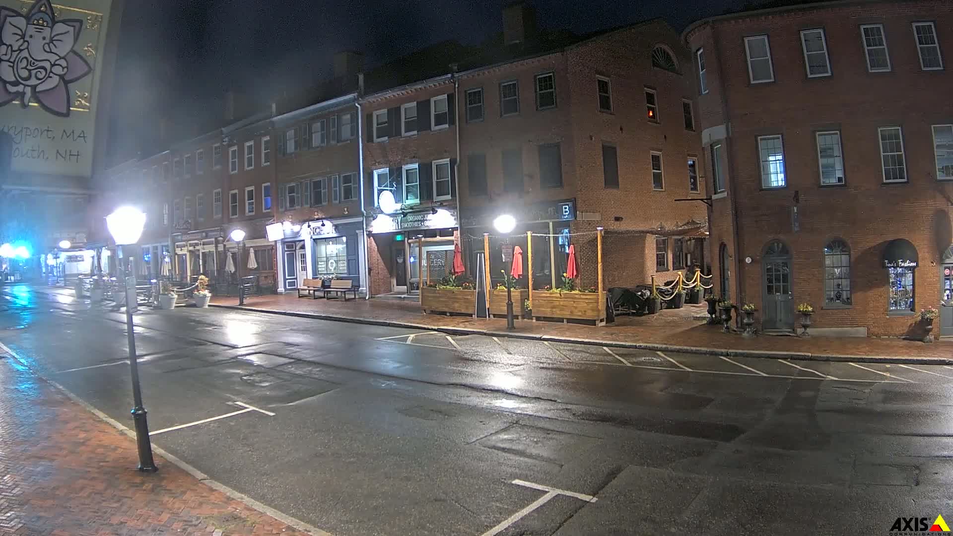 A wet, nighttime street scene shows several brick buildings with outdoor seating areas, lit by streetlights.