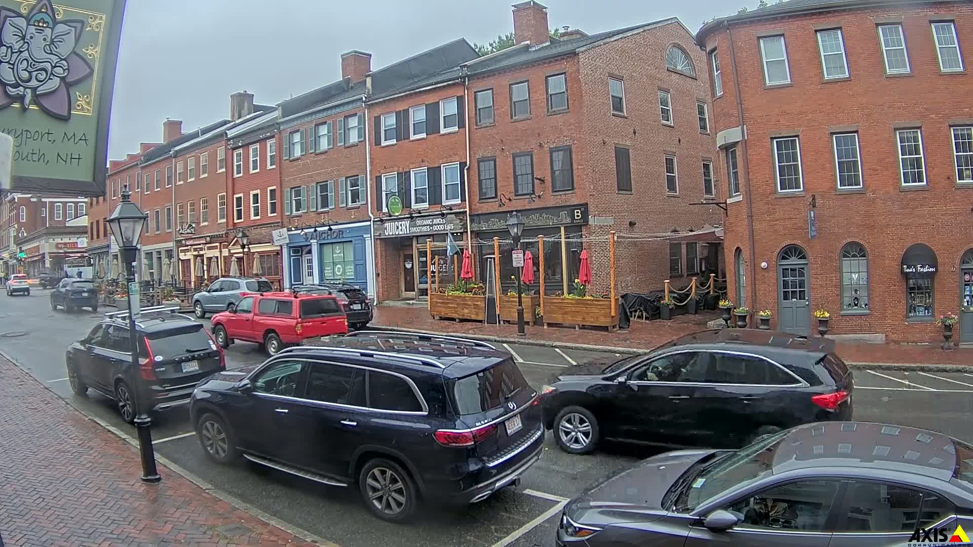 A rain-slicked street lined with brick buildings and parked cars, featuring outdoor seating at a restaurant.