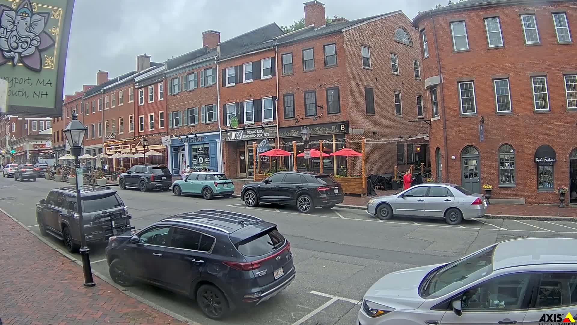 A rain-slicked street lined with brick buildings and parked cars, featuring outdoor seating at a restaurant.