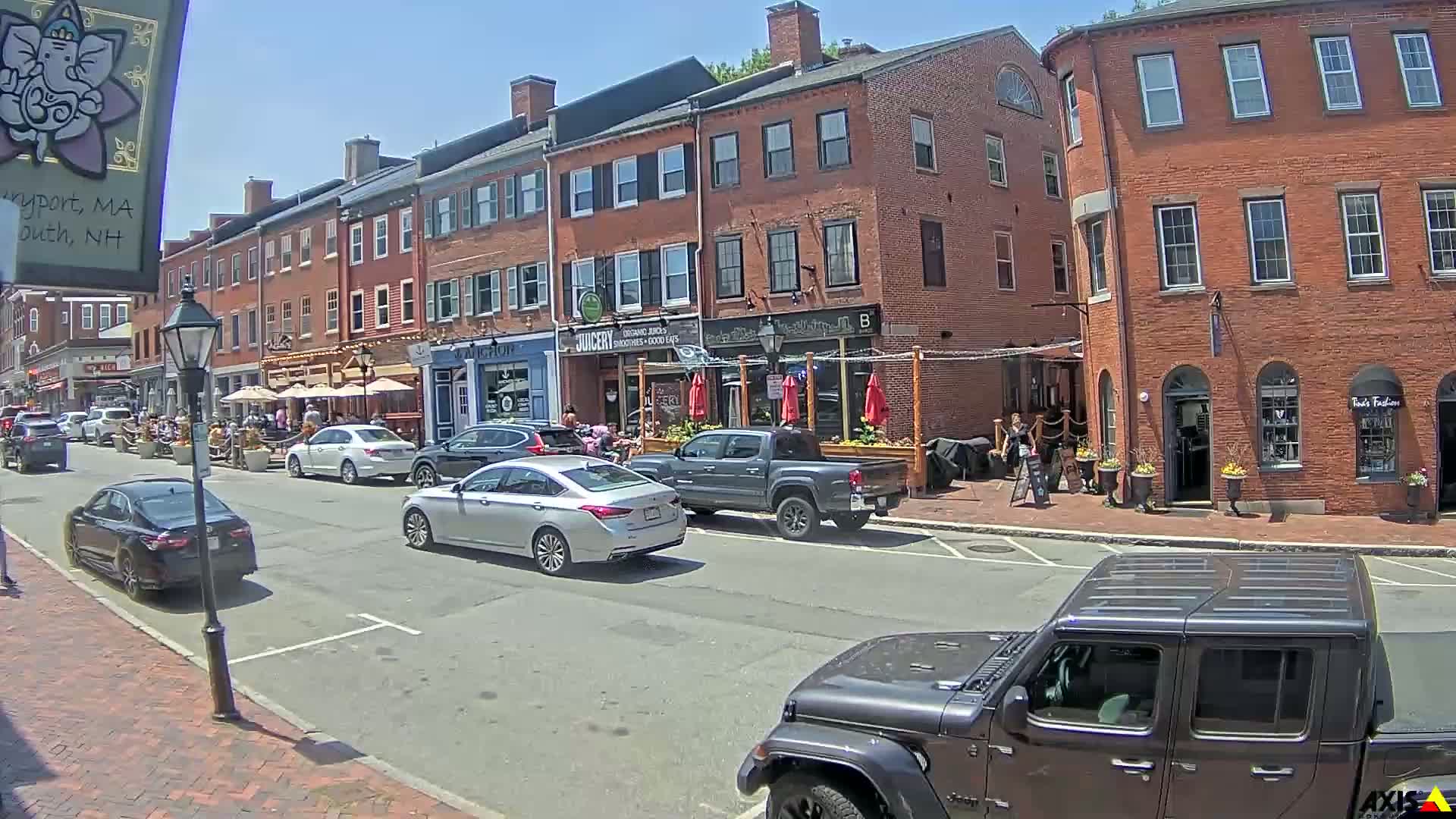 A sunny street scene shows several brick buildings with outdoor seating areas, cars parked along the curb, and people walking along the sidewalk.