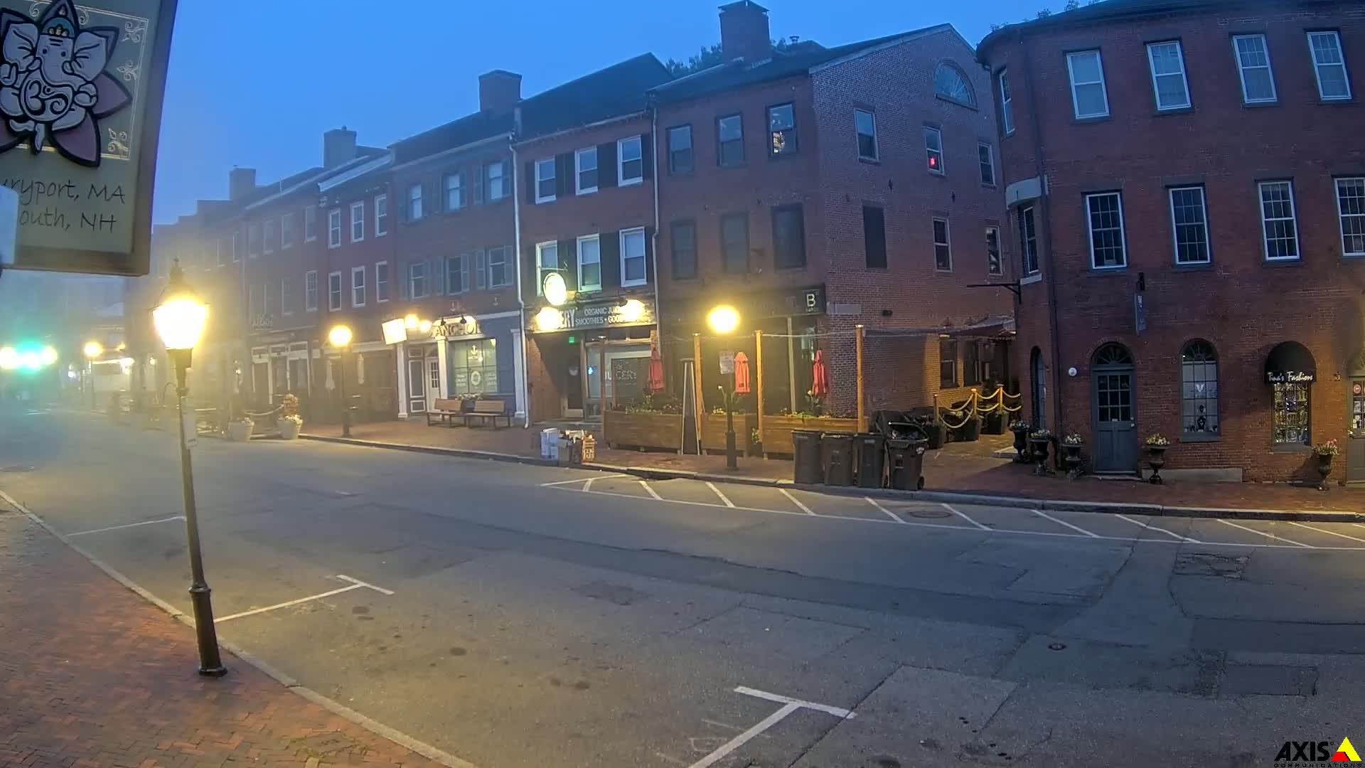 A foggy, twilight street scene shows several brick buildings with shops and outdoor seating areas, lit by street lamps.