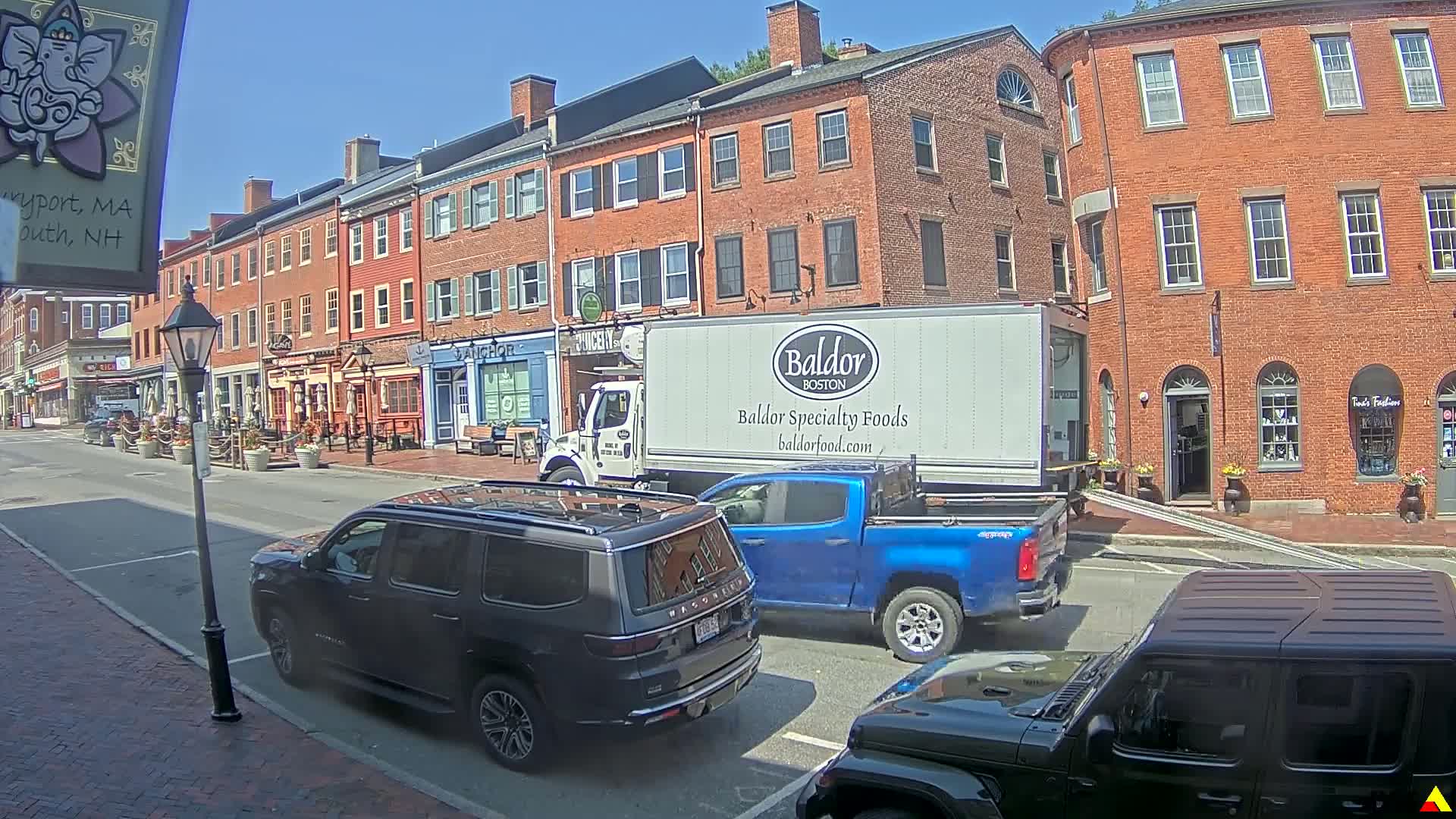 A sunny day on a brick-lined street with several shops and parked cars, including a dark-colored SUV, a blue pickup truck, and a delivery truck.
