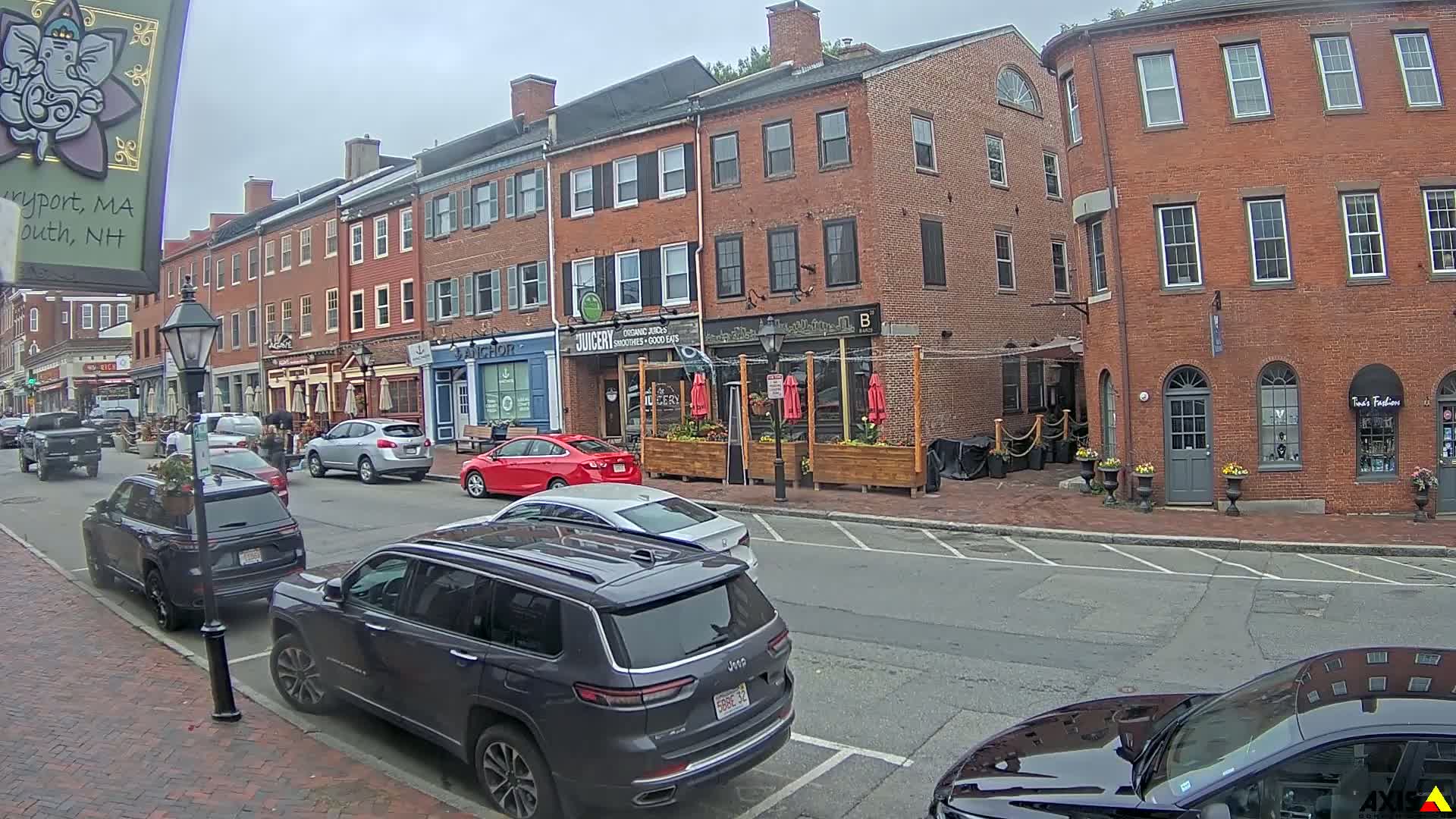 A street scene on an overcast day shows several parked cars in front of brick buildings with various shops and restaurants.