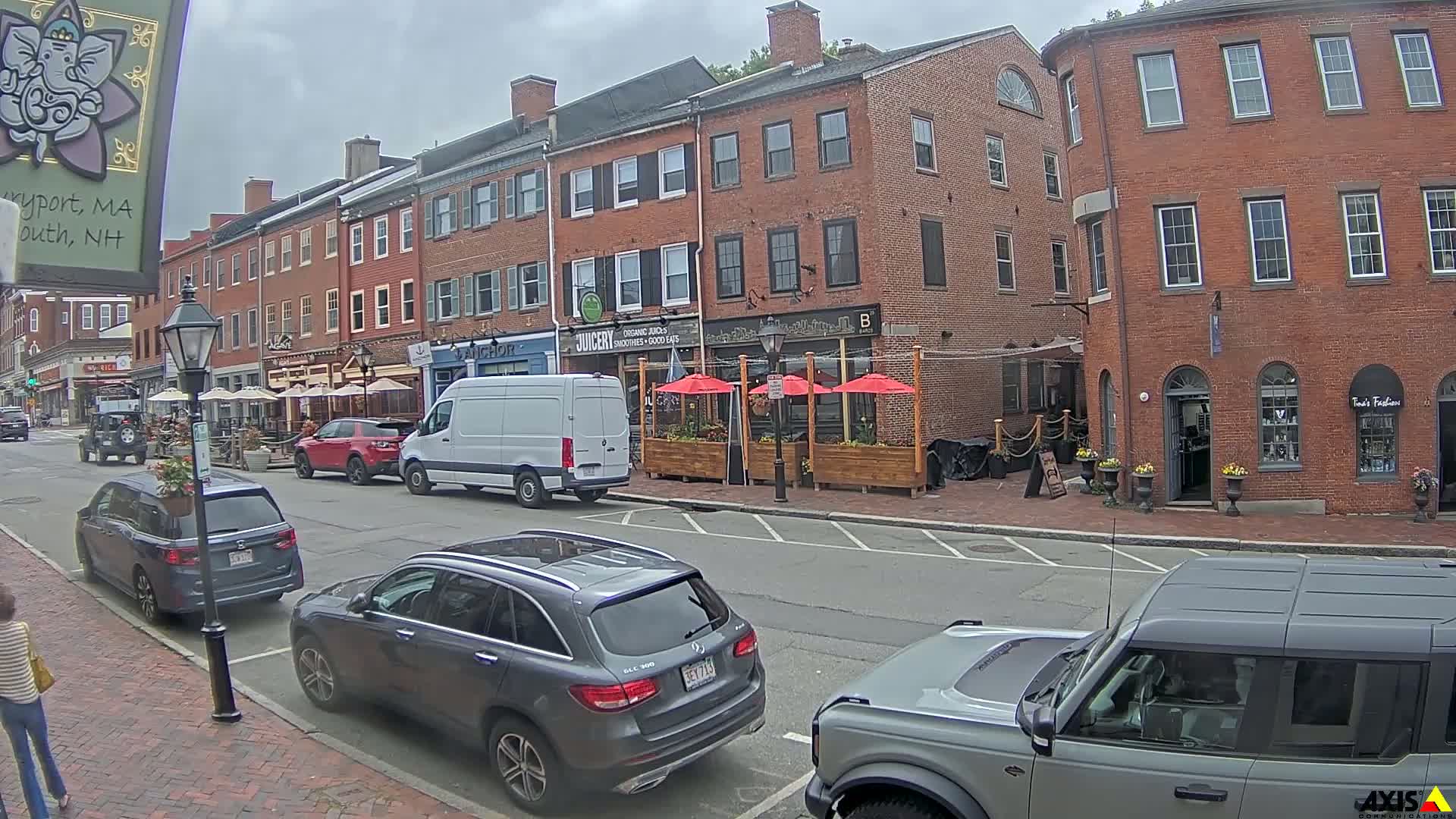 A street scene on an overcast day shows several parked cars in front of brick buildings with outdoor seating areas.