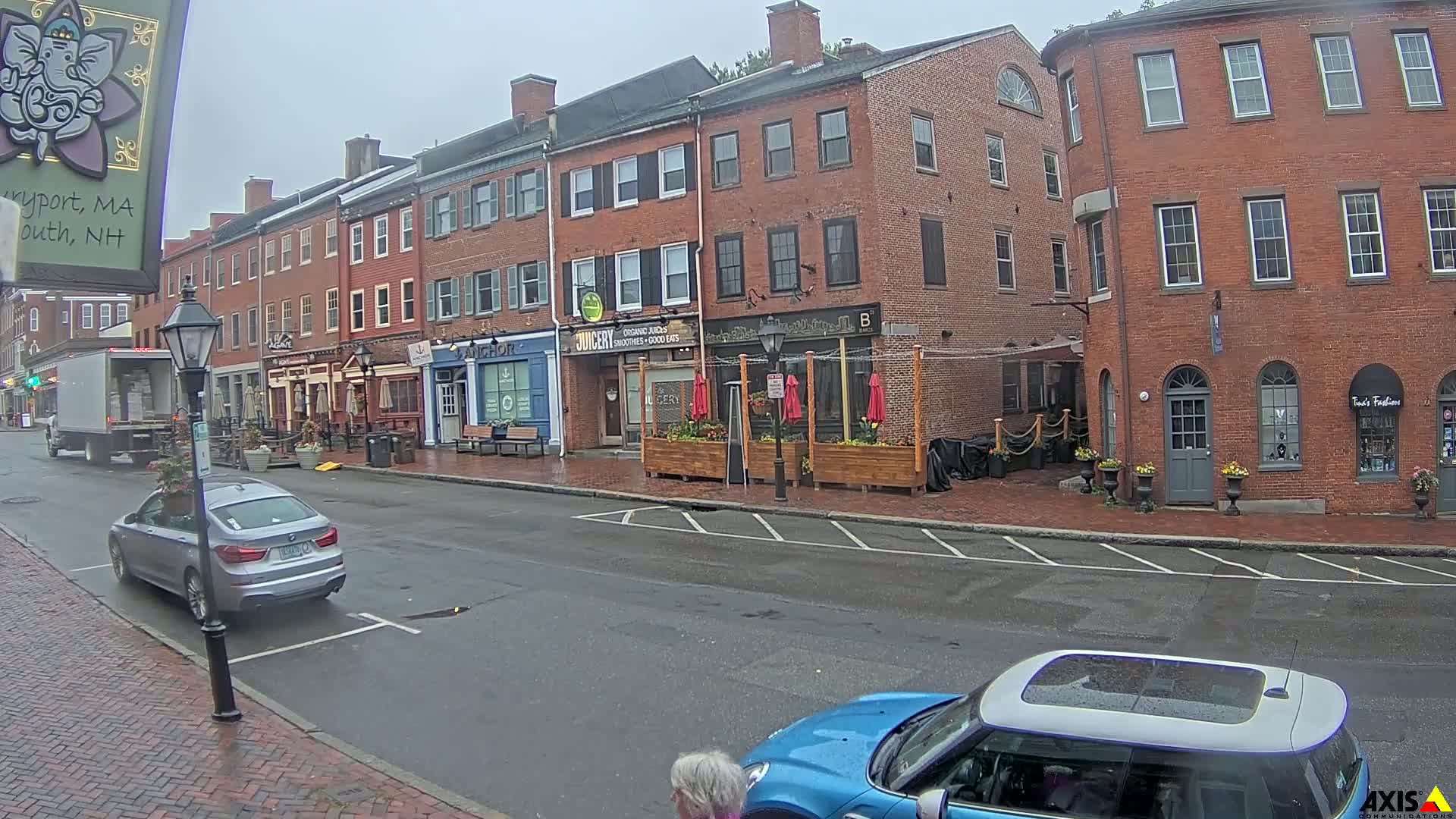 A wet city street scene shows brick buildings with various shops and restaurants, a parked silver car, and a blue car partially visible in the foreground, under an overcast sky.