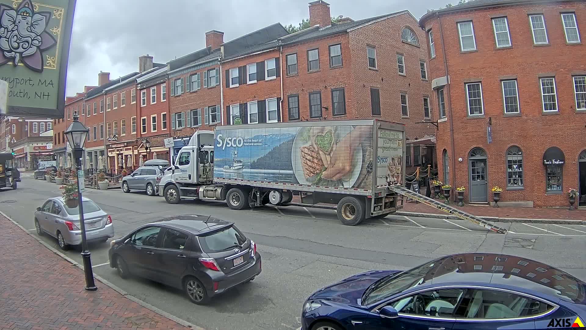 A Sysco delivery truck is parked on a city street with overcast skies, near several parked cars and brick buildings.