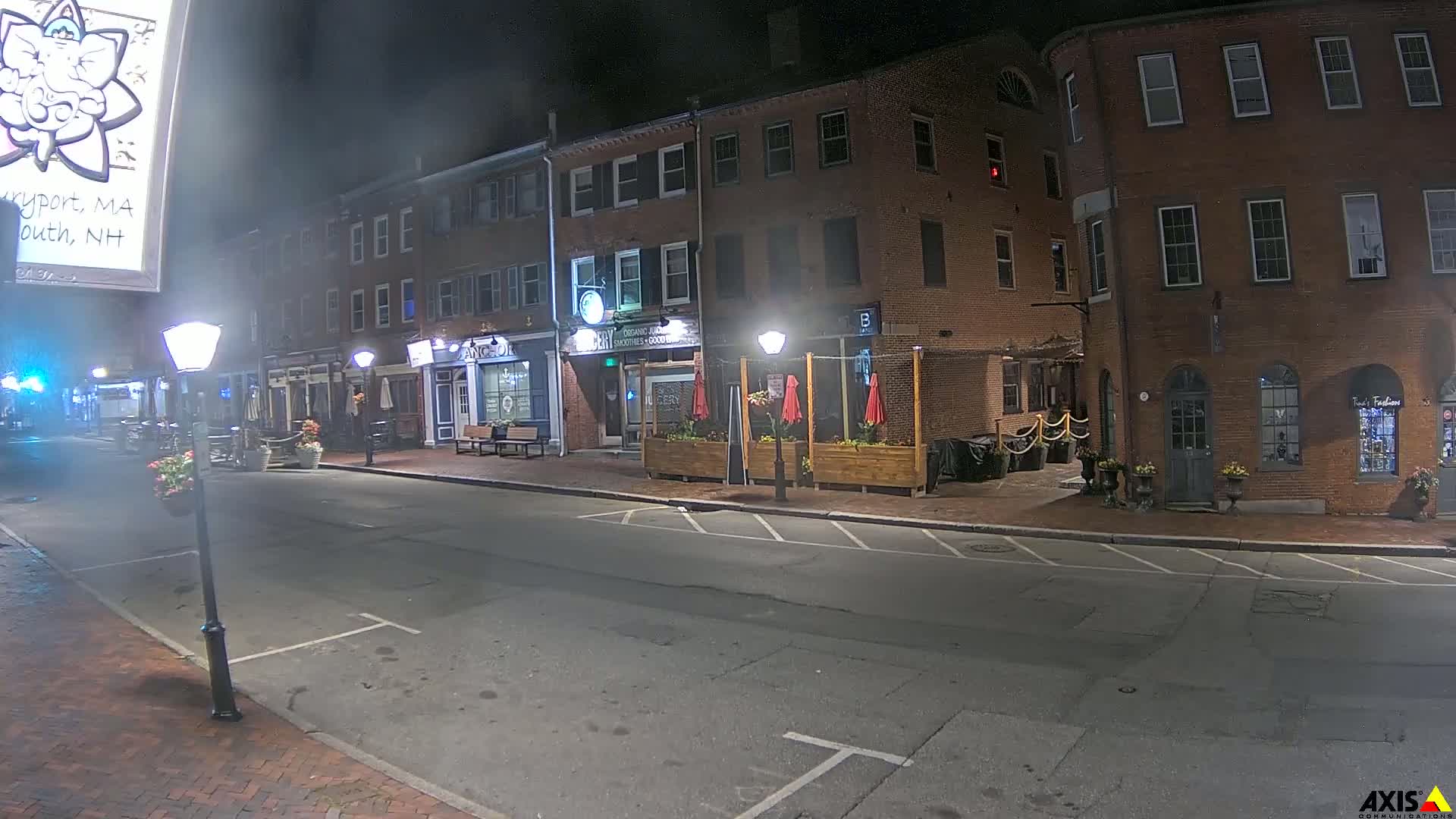 A nighttime street scene shows several brick buildings with outdoor seating areas, lit by street lamps under a dark sky.