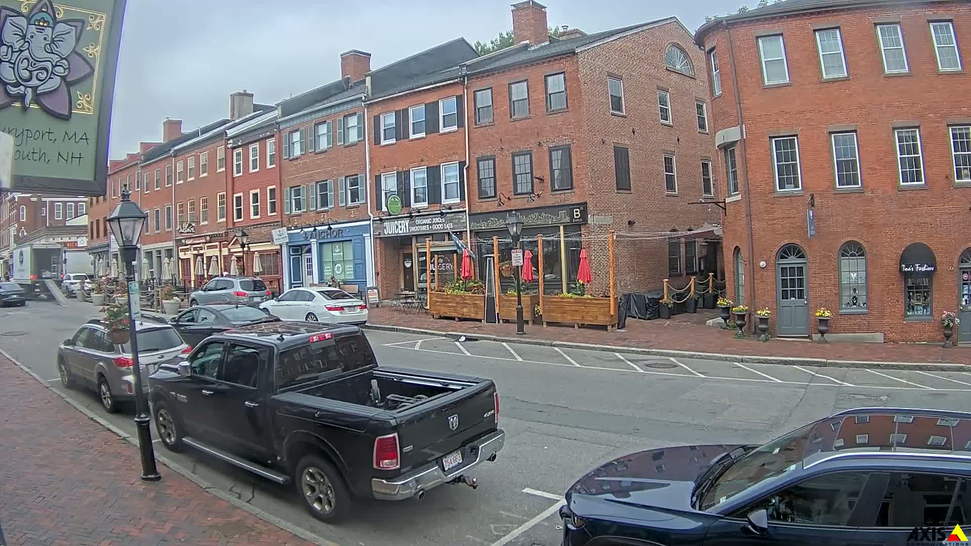 A partly cloudy day on a brick-lined street with parked cars and several businesses, including a juice bar and a clothing store.