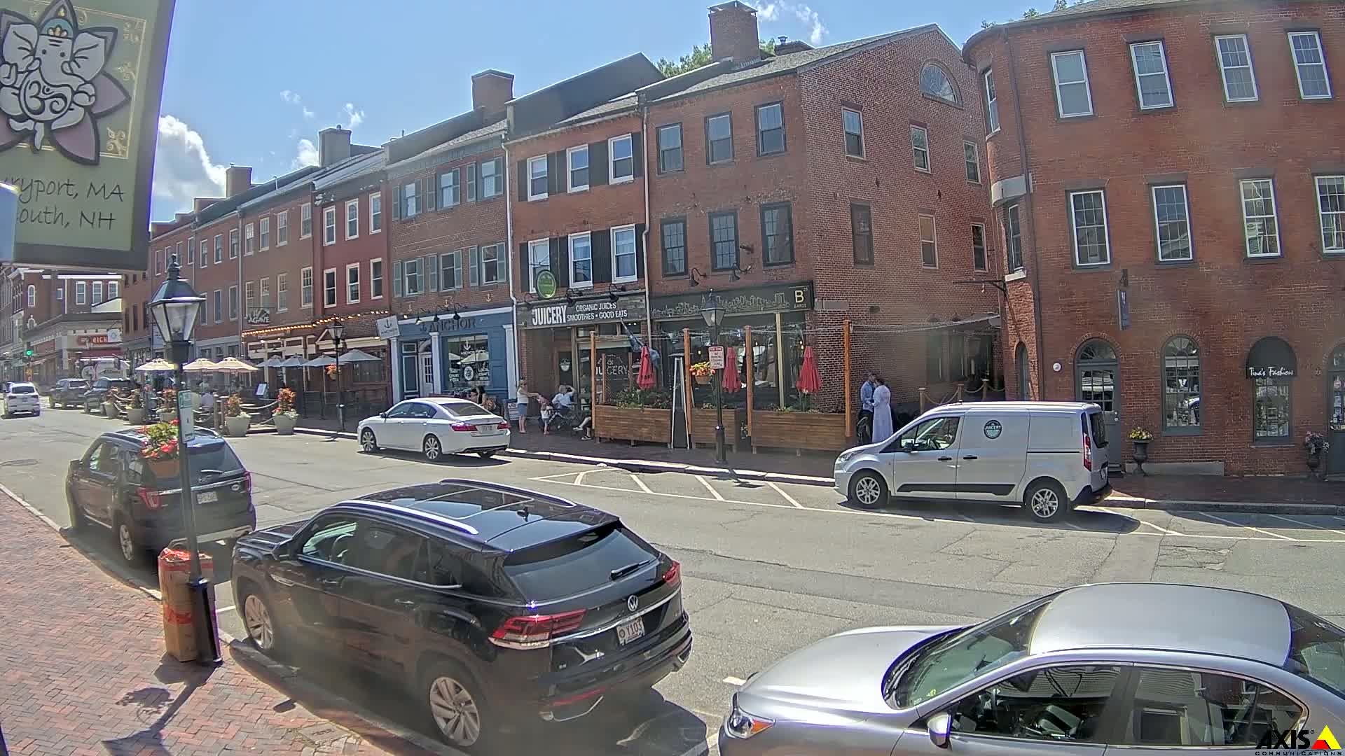 A sunny day on a brick-lined street with parked cars, pedestrians, and outdoor seating at various businesses.