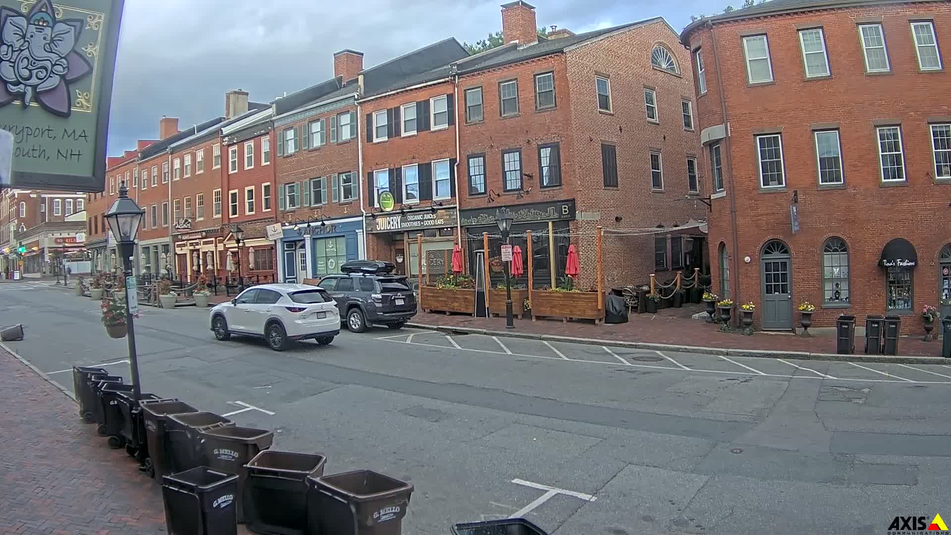 A street scene on an overcast day shows several brick buildings with outdoor seating areas and parked cars, along with trash cans lining the curb.