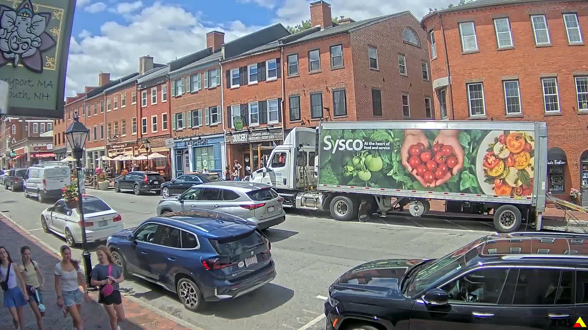 A sunny day on a city street with brick buildings, restaurants with outdoor seating, parked cars, and a Sysco delivery truck.