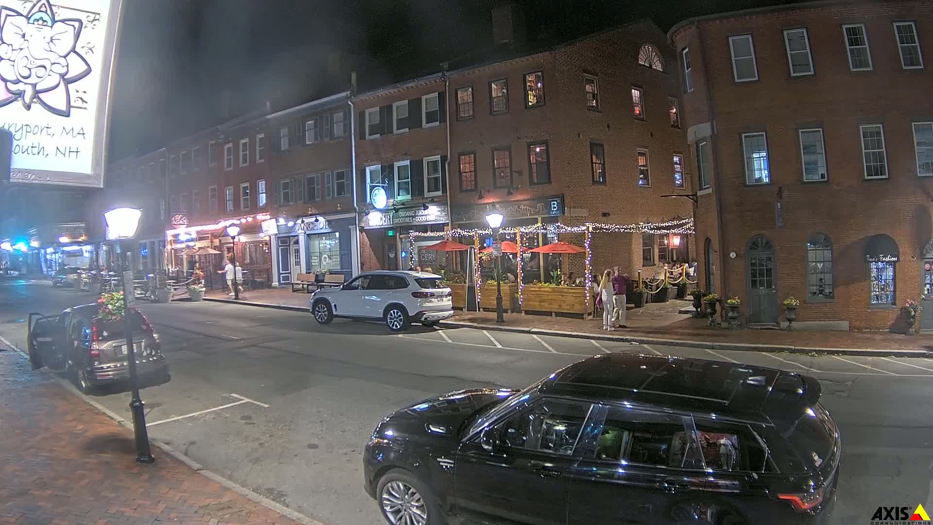 At night, a street with brick buildings and outdoor restaurant seating is illuminated, with cars parked along the curb and a few pedestrians visible.
