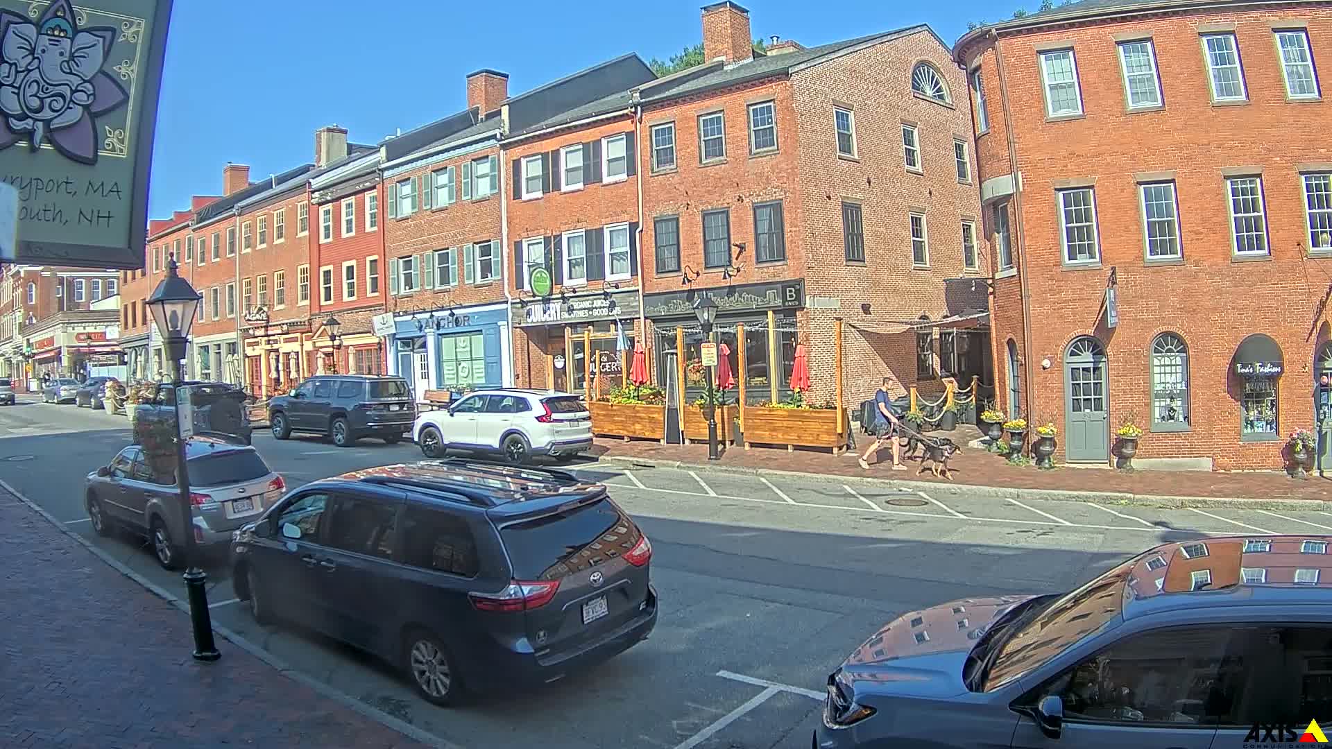 A sunny street scene shows several parked cars in front of brick buildings with shops and restaurants on a clear day.