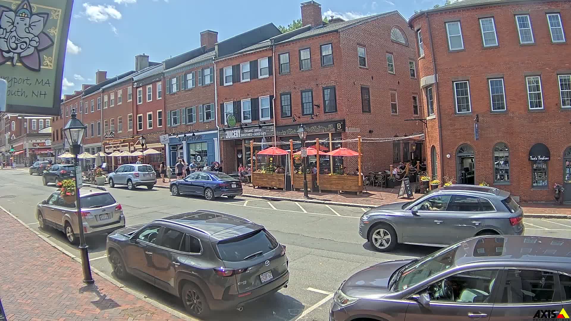 A sunny day on a brick-lined street with parked cars and outdoor seating at several restaurants.