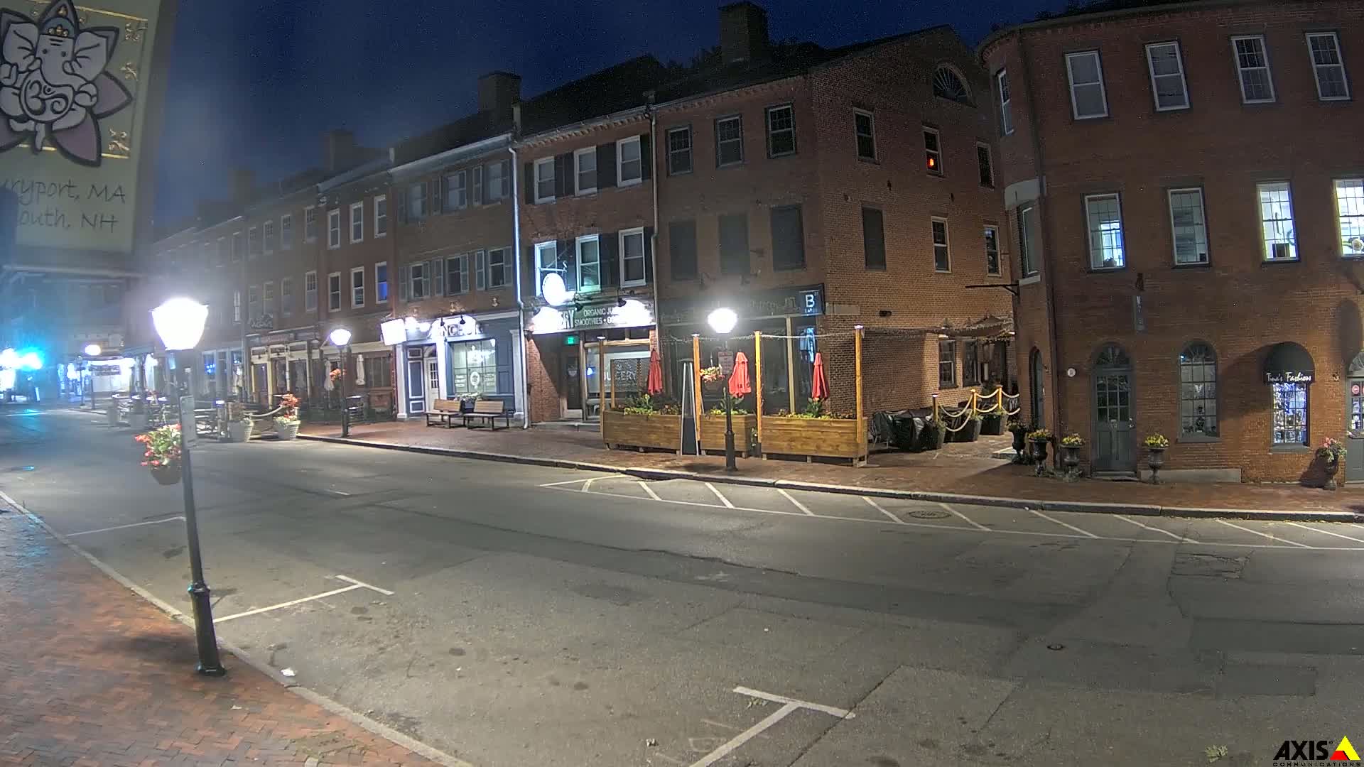 A nighttime street scene shows several brick buildings with outdoor seating areas and streetlights illuminating a mostly empty road.