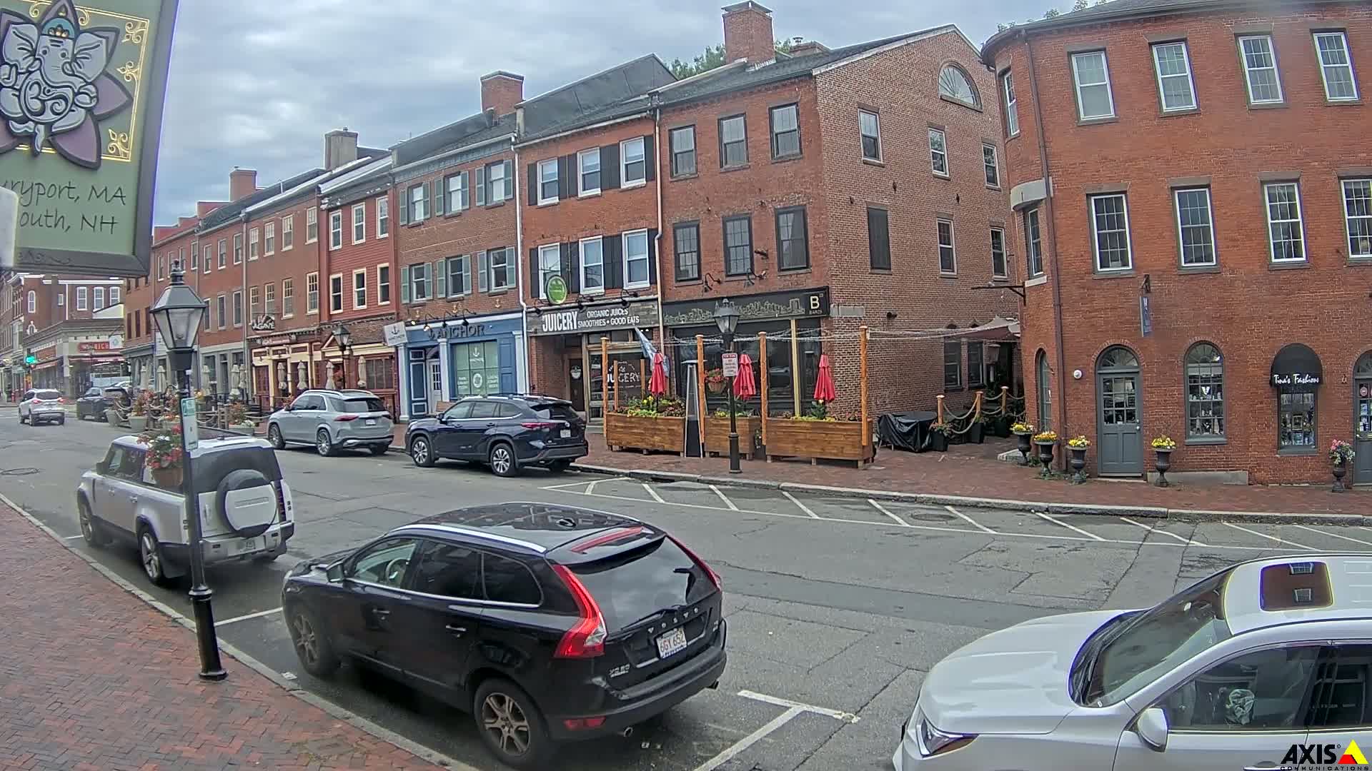 A street scene on an overcast day shows several parked cars in front of brick buildings with various shops and restaurants, including one with outdoor seating.