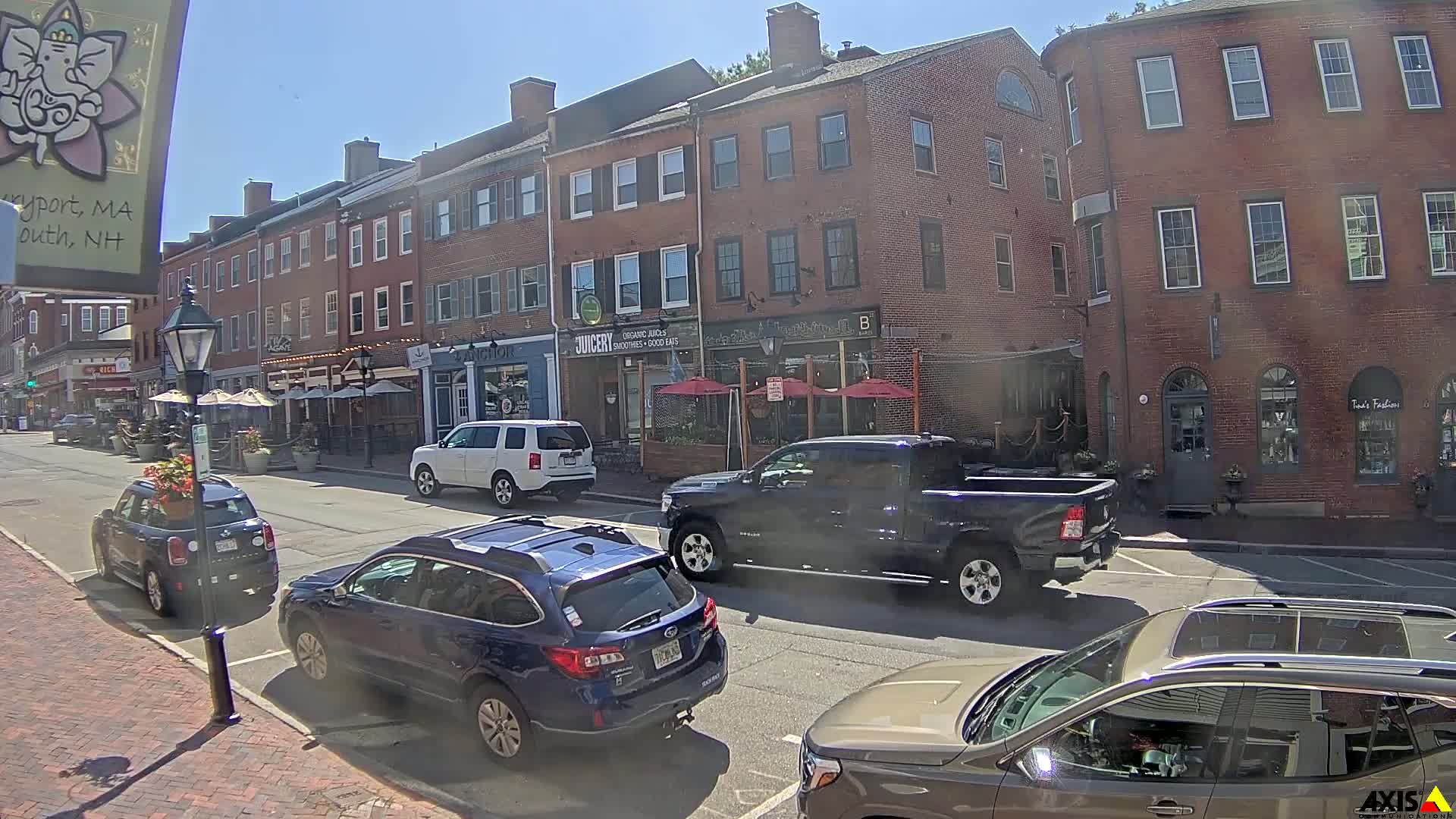 A sunny street scene shows several cars parked and driving past brick buildings with outdoor seating areas in front of various businesses.