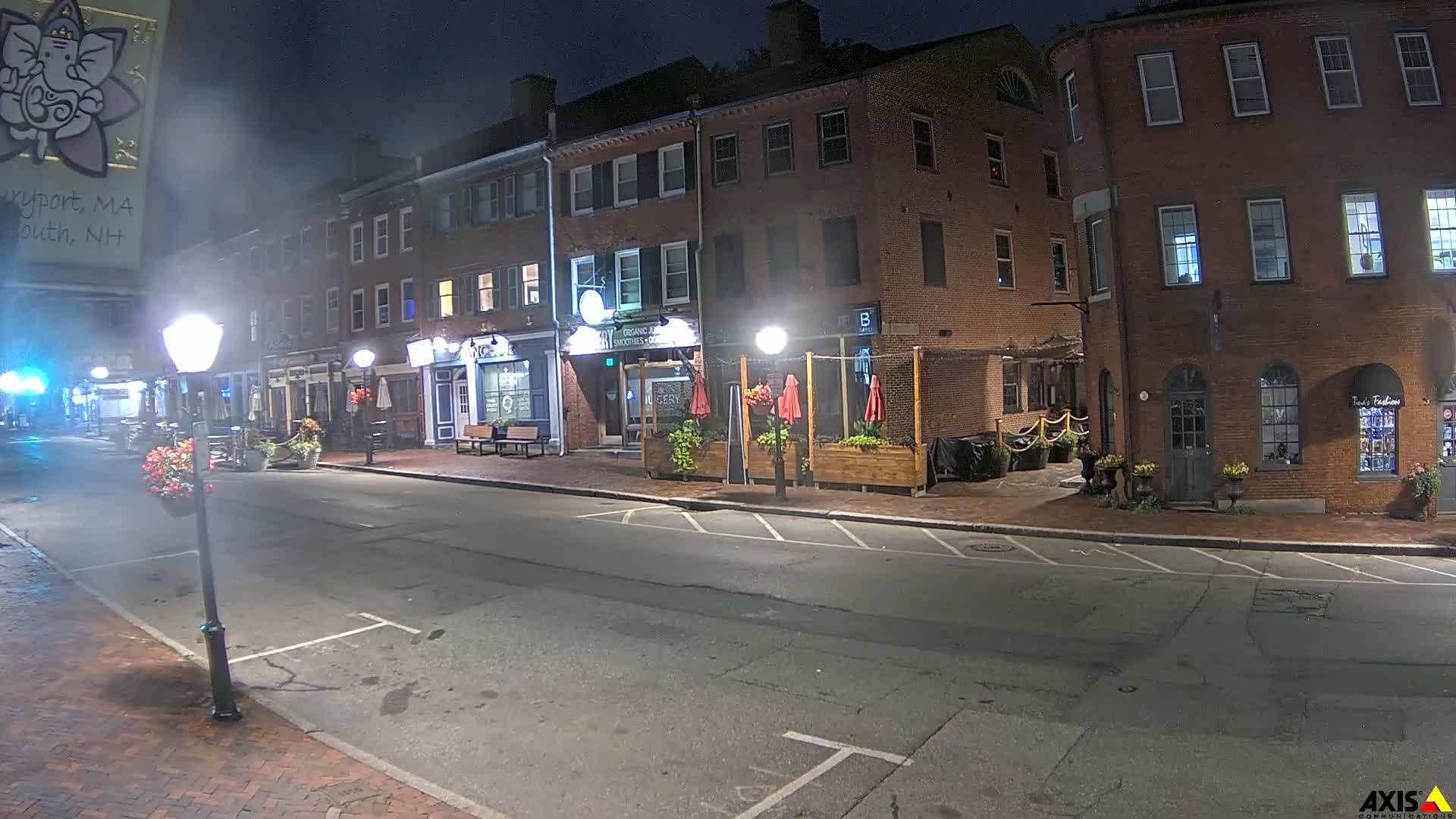 A quiet, nighttime street scene shows brick buildings with outdoor seating areas and lampposts lining a mostly empty road.