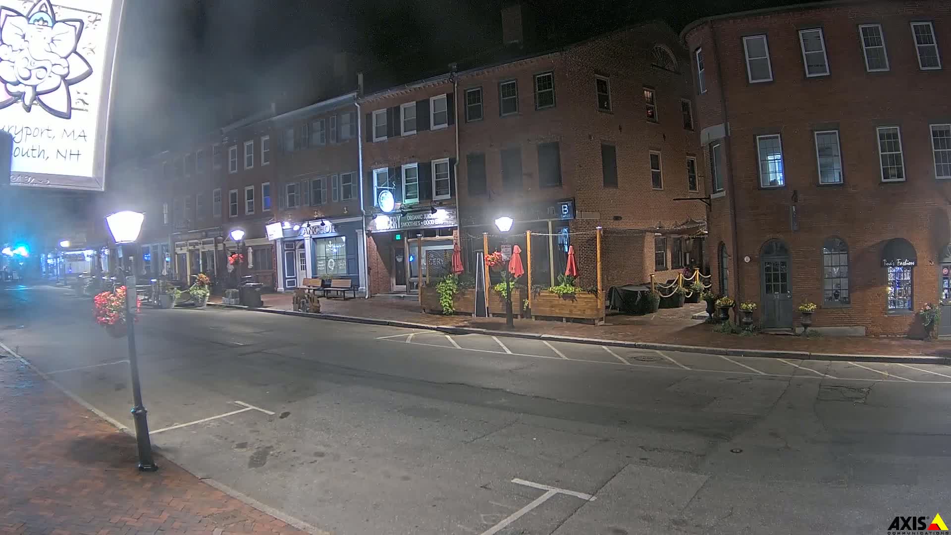 A quiet, empty street at night lined with brick buildings, some with outdoor seating areas, under dark, clear skies.