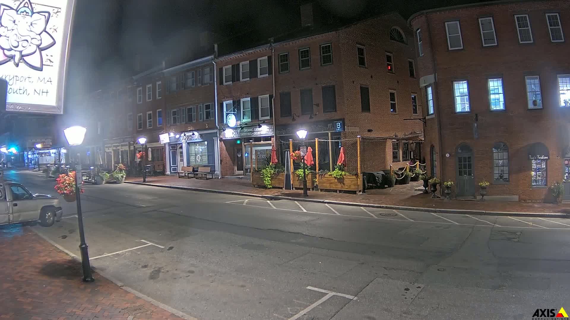 A nighttime street scene shows a brick building with businesses on the ground floor, outdoor seating with red umbrellas, and a few parked cars.