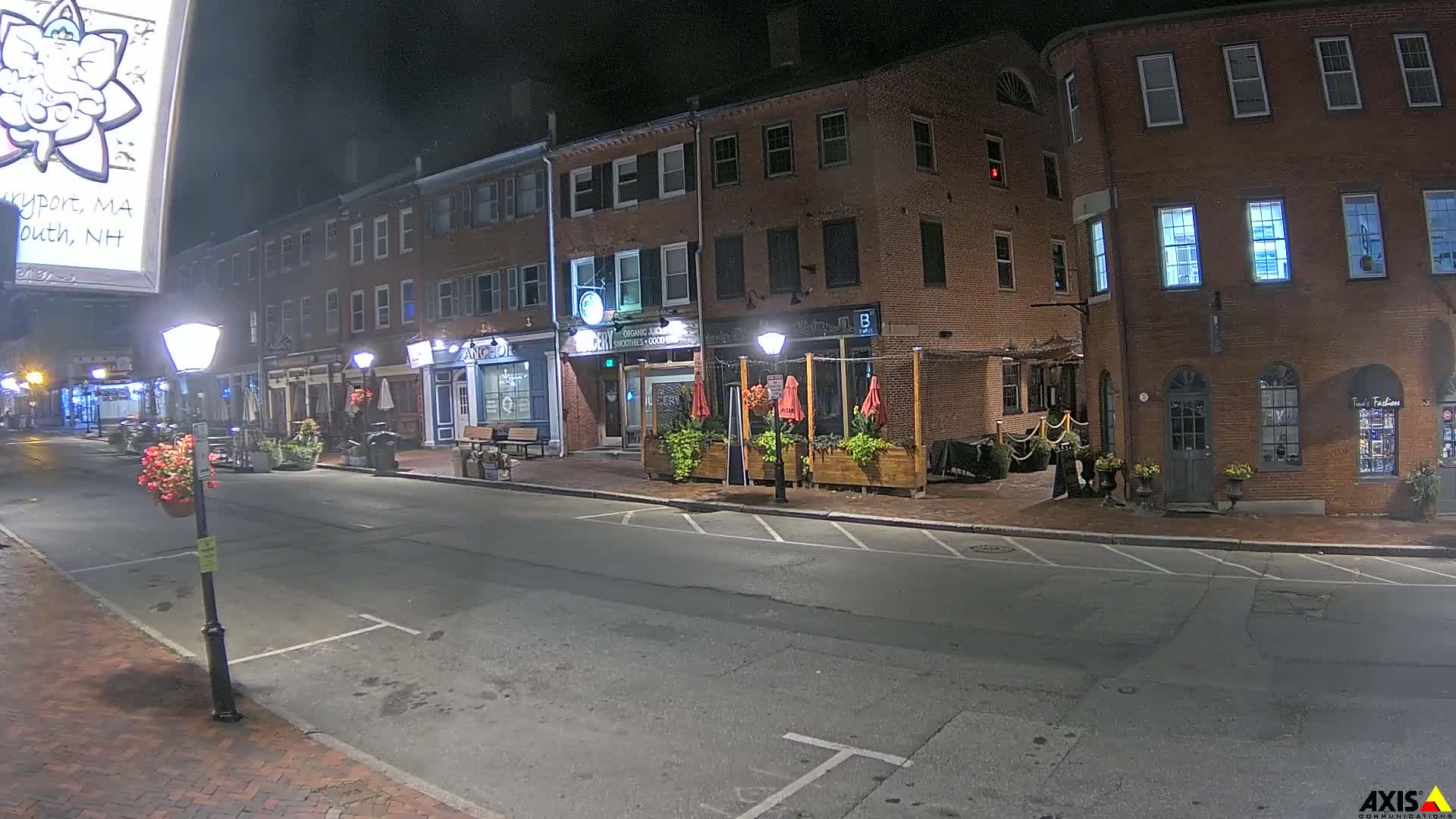 A nighttime street scene shows a brick building with businesses on the ground floor, outdoor seating with red umbrellas, and a few parked cars.