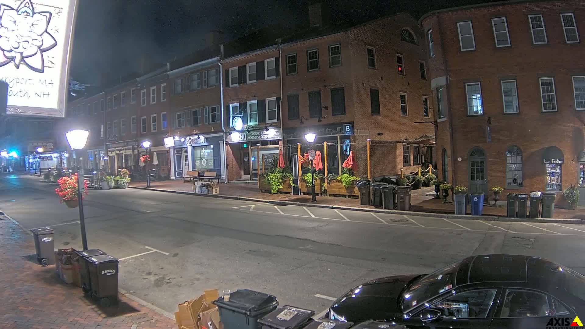 A nighttime street scene shows a mostly empty road lined with brick buildings, some with outdoor seating areas, under clear skies.