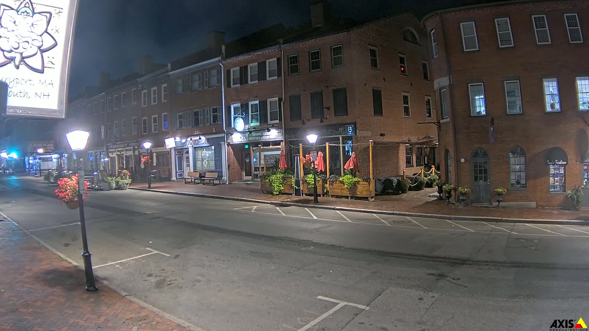 A nighttime street scene shows a row of brick buildings with outdoor seating areas, illuminated by streetlights, under a dark sky.