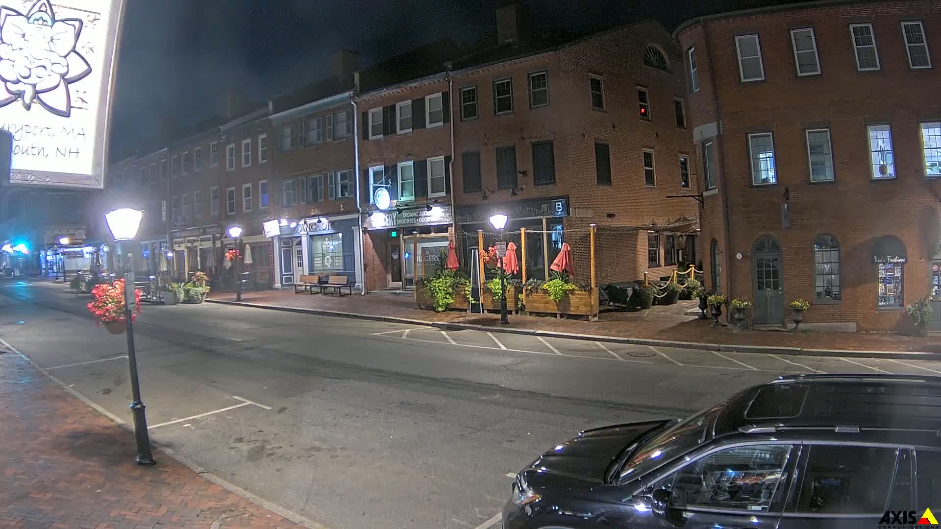 A nighttime street scene shows a mostly empty road lined with brick buildings, some with outdoor seating areas, lit by street lamps.
