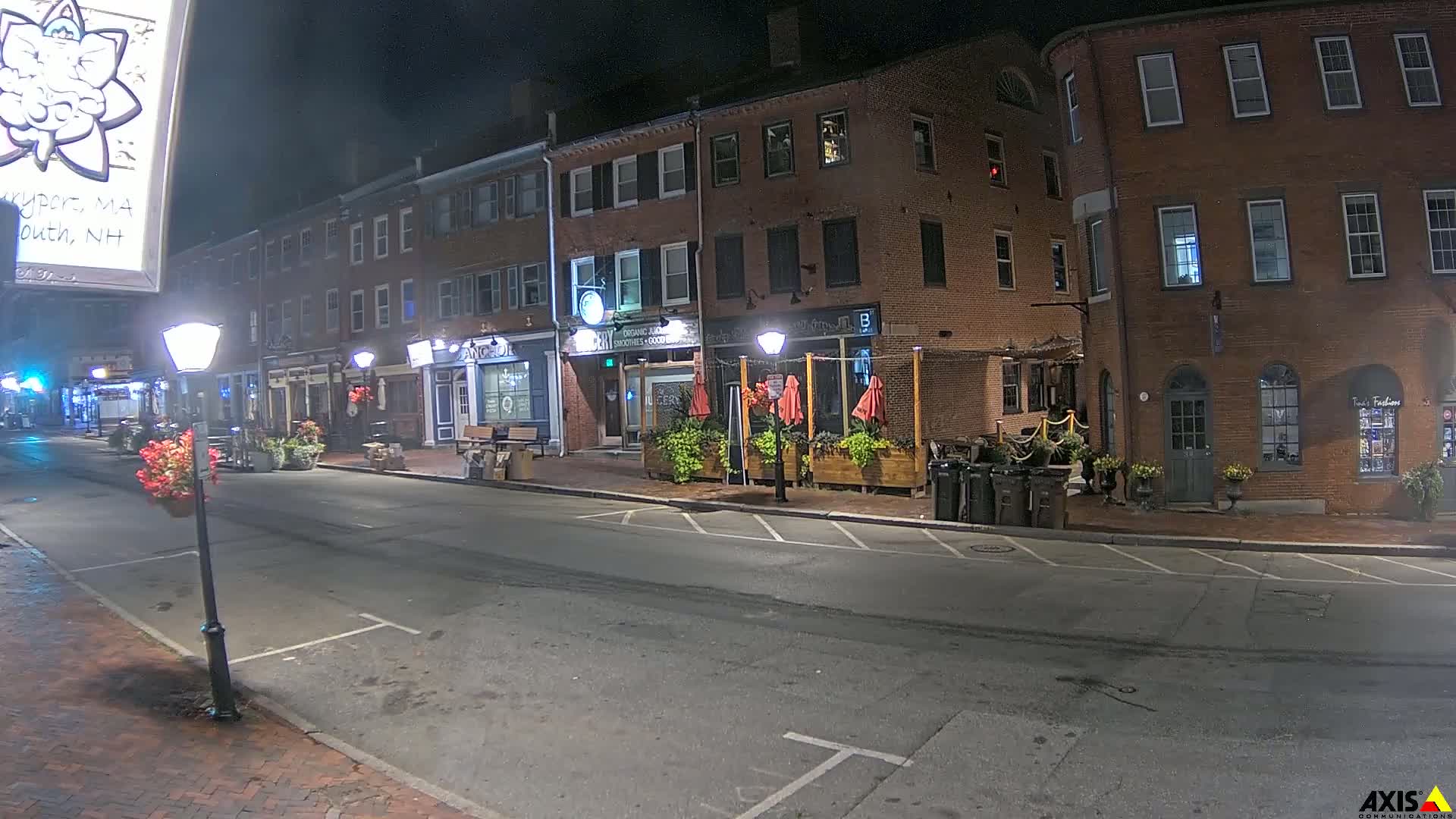 A mostly empty street at night lined with brick buildings, some with outdoor seating areas, under dark skies.