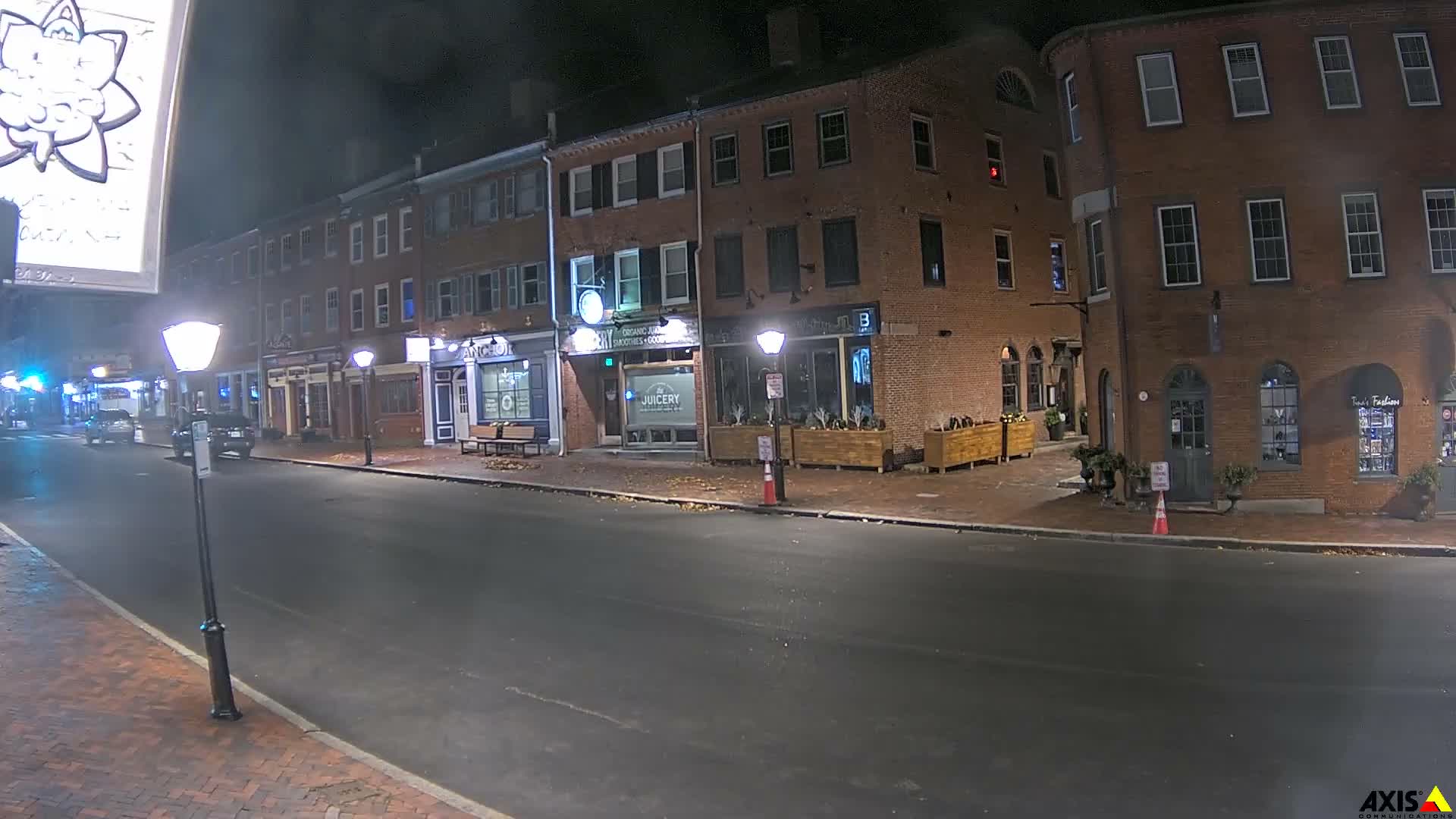 A mostly empty street at night lined with brick buildings, some with outdoor seating areas, under dark skies.