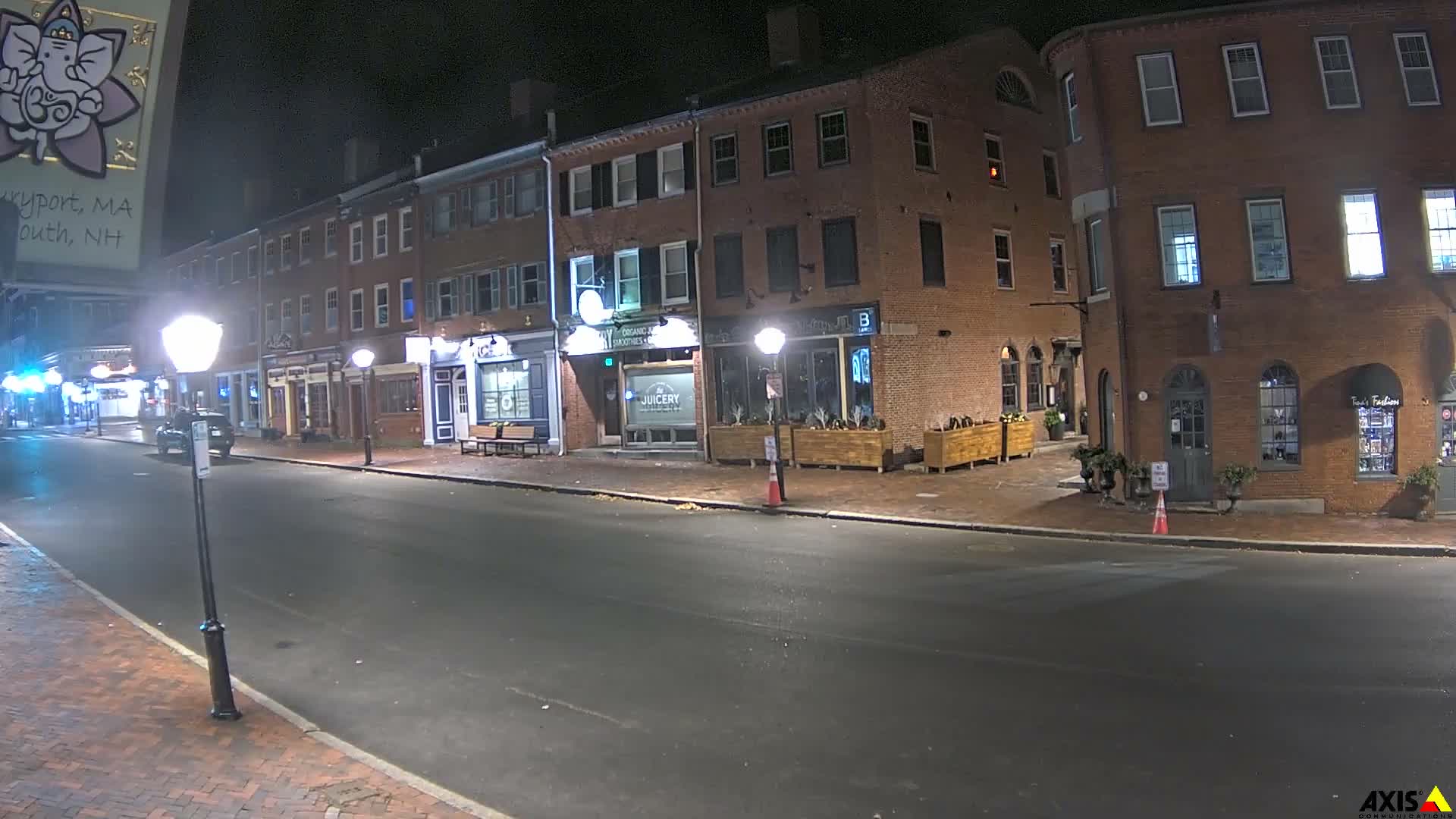 A quaint brick-lined street at night, illuminated by streetlights and glowing shop windows, appears quiet and dry under a clear sky.