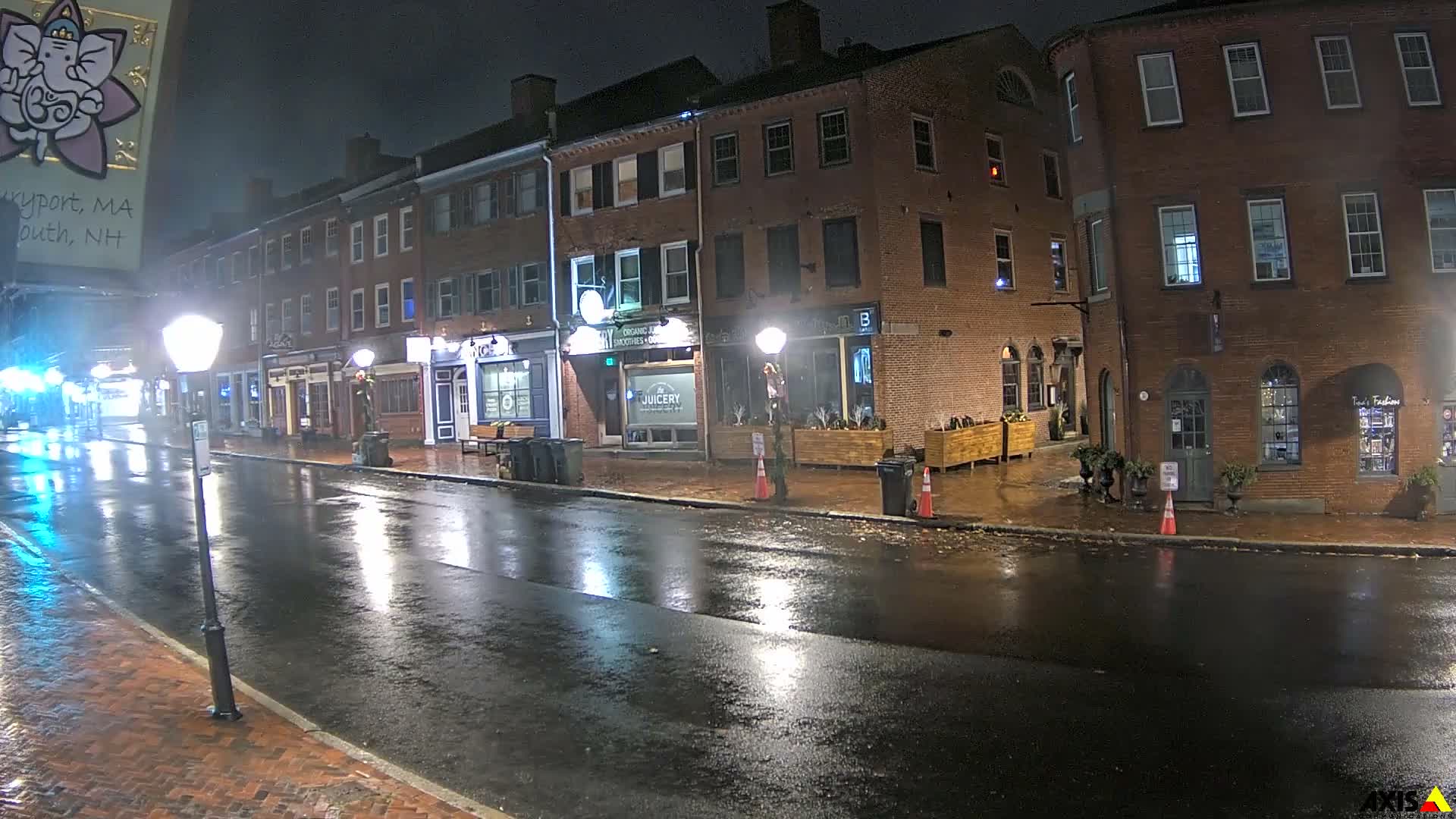 A wet, reflective street runs alongside historic brick buildings with illuminated shopfronts on a rainy night.