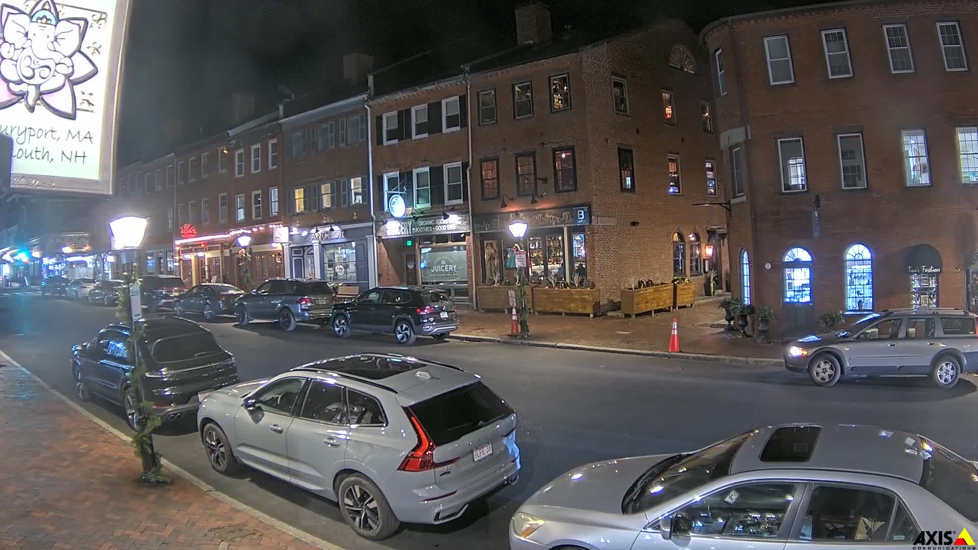 A clear night reveals a bustling street lined with historic brick buildings housing various shops, illuminated by streetlights and storefront glow, with numerous cars parked and moving along the roadway.