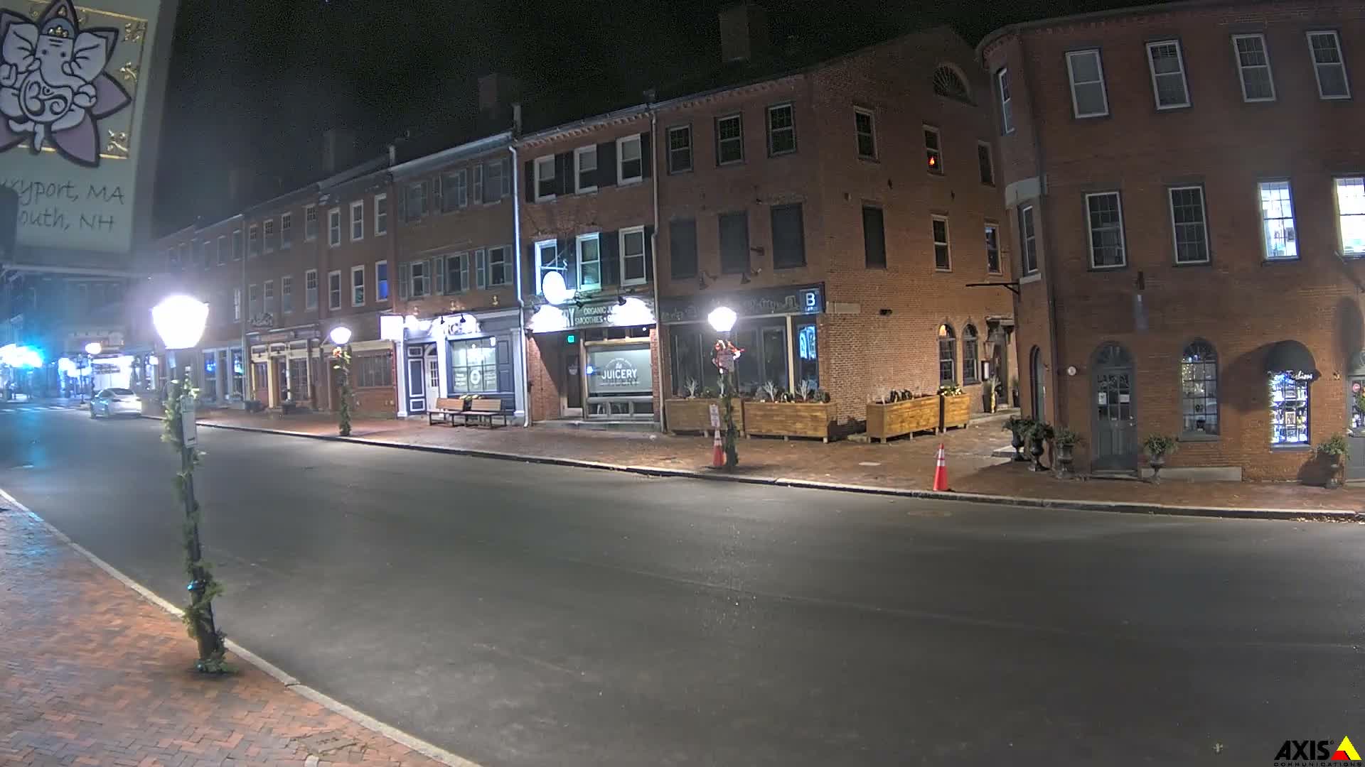 A quiet street at night is illuminated by streetlights and glowing shopfronts along multi-story brick buildings, revealing an empty road and brick sidewalks, with some lampposts adorned with greenery under clear skies.