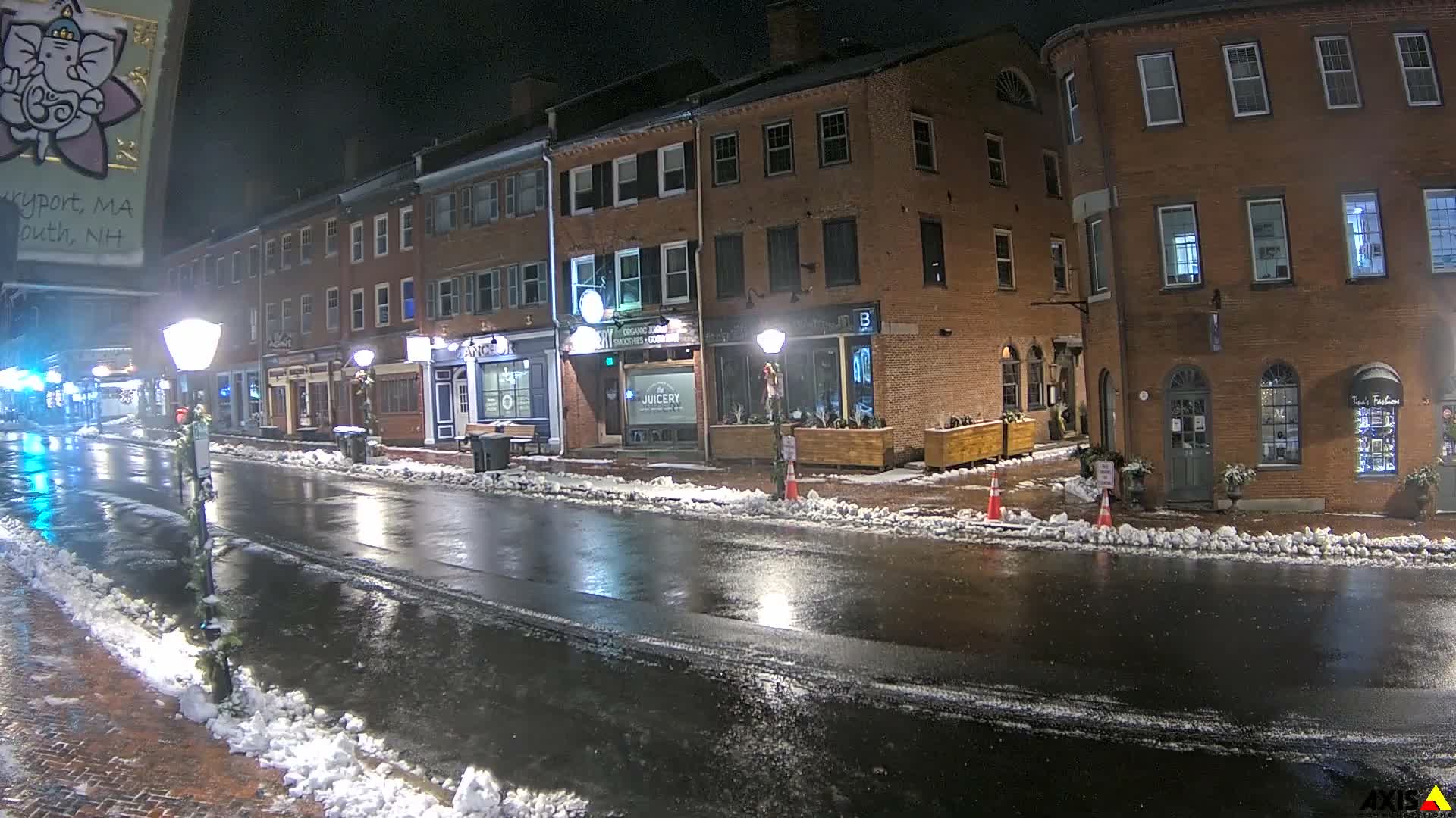 A nighttime view reveals a wet city street reflecting streetlights, flanked by historic brick buildings, with patches of snow remaining along the curbs.