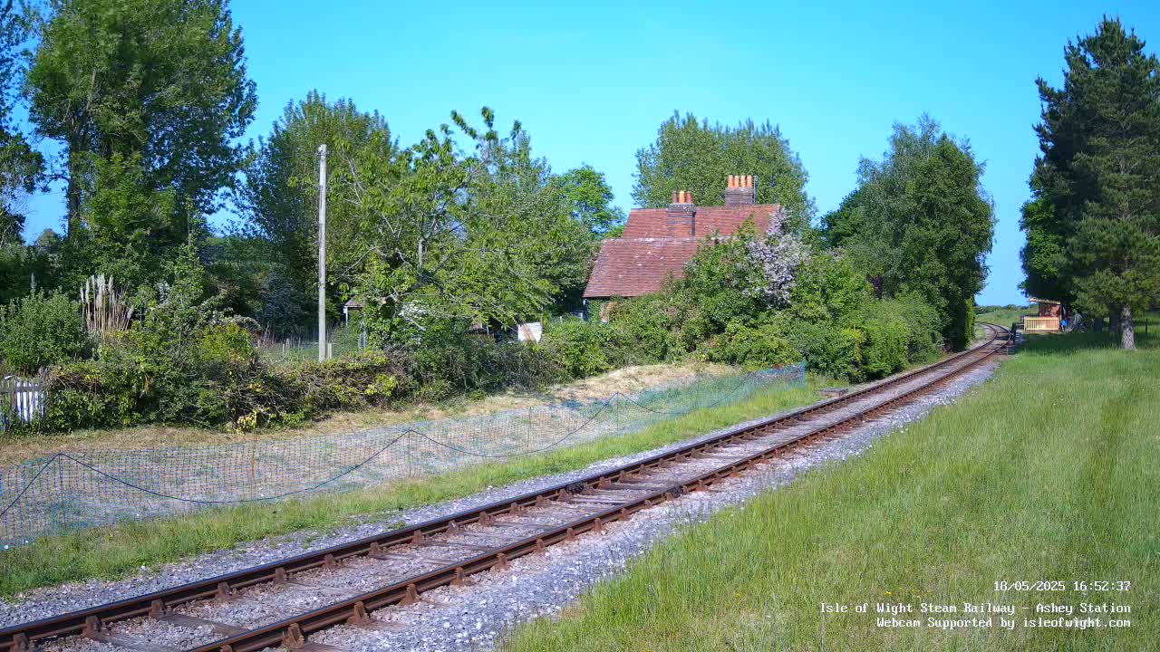 A train track winds through a grassy field toward a small, red-roofed building nestled amongst green trees under a clear blue sky.