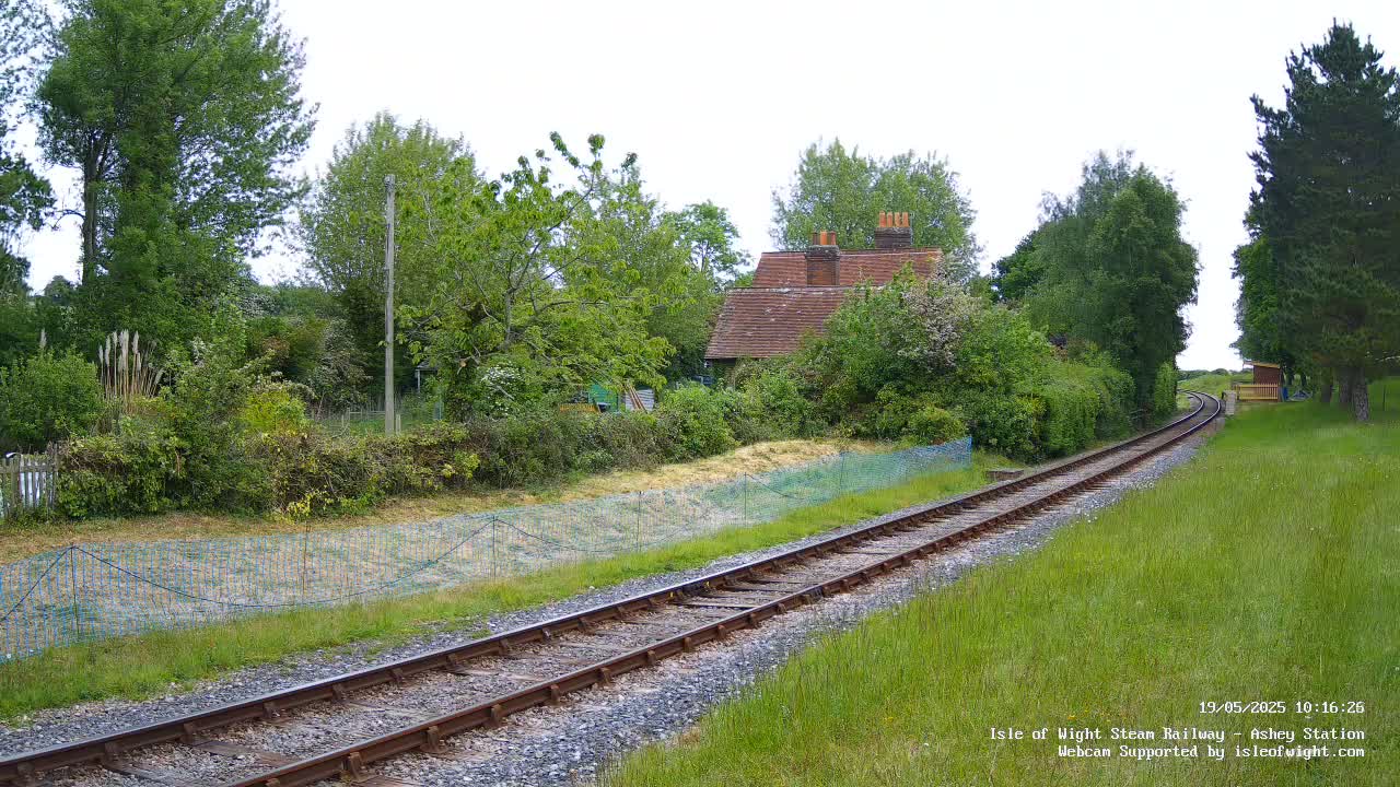 A train track extends from the foreground into the distance, passing by a small cottage nestled among lush green vegetation under a partly cloudy sky.