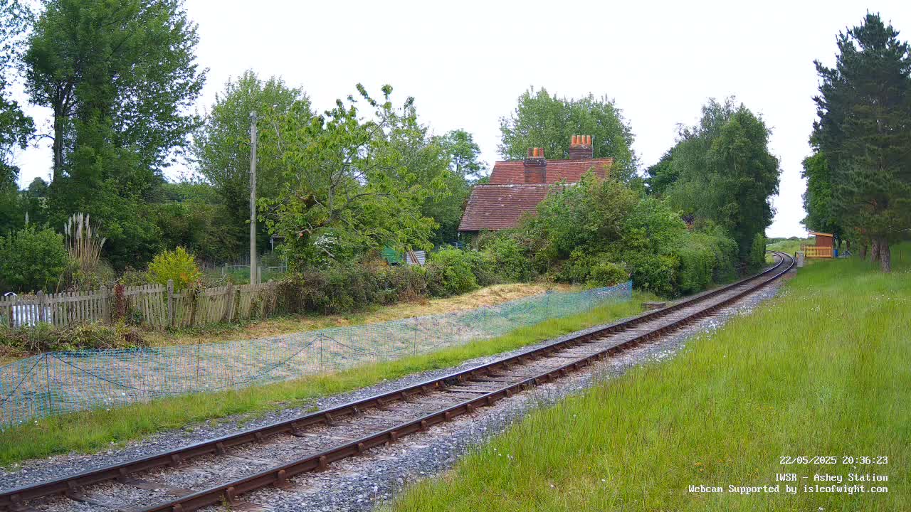 A railway track curves gently past a small brick house nestled among green trees and overgrown vegetation on a partly cloudy day.