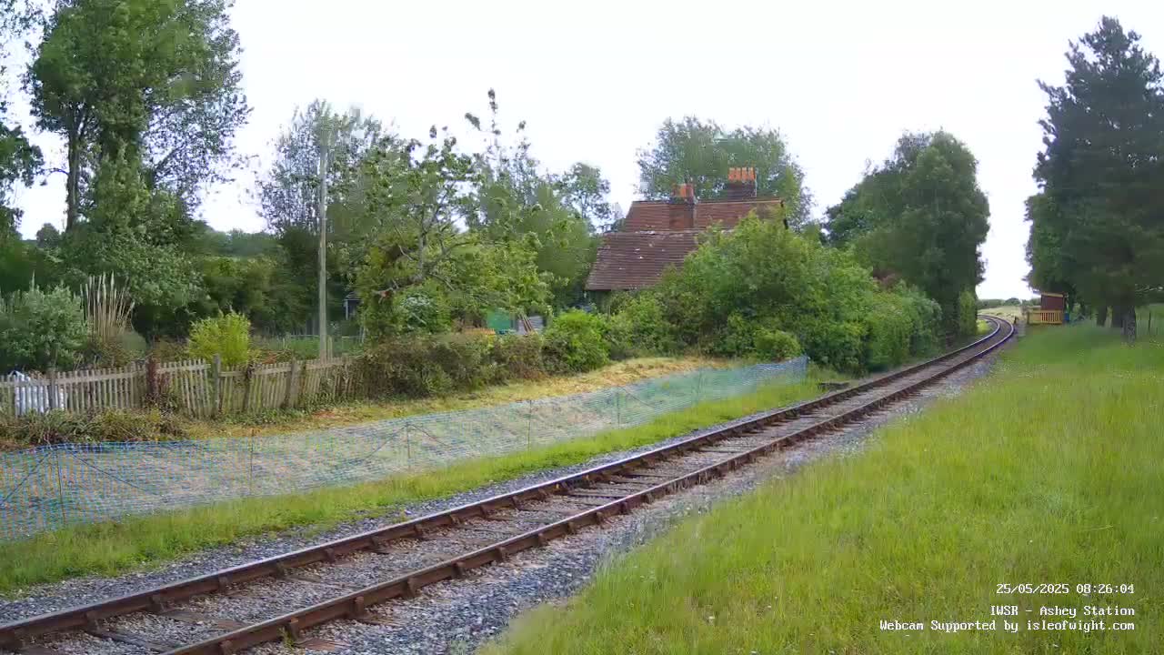 A railway track winds through a verdant landscape, passing a small house nestled amongst trees on an overcast day.