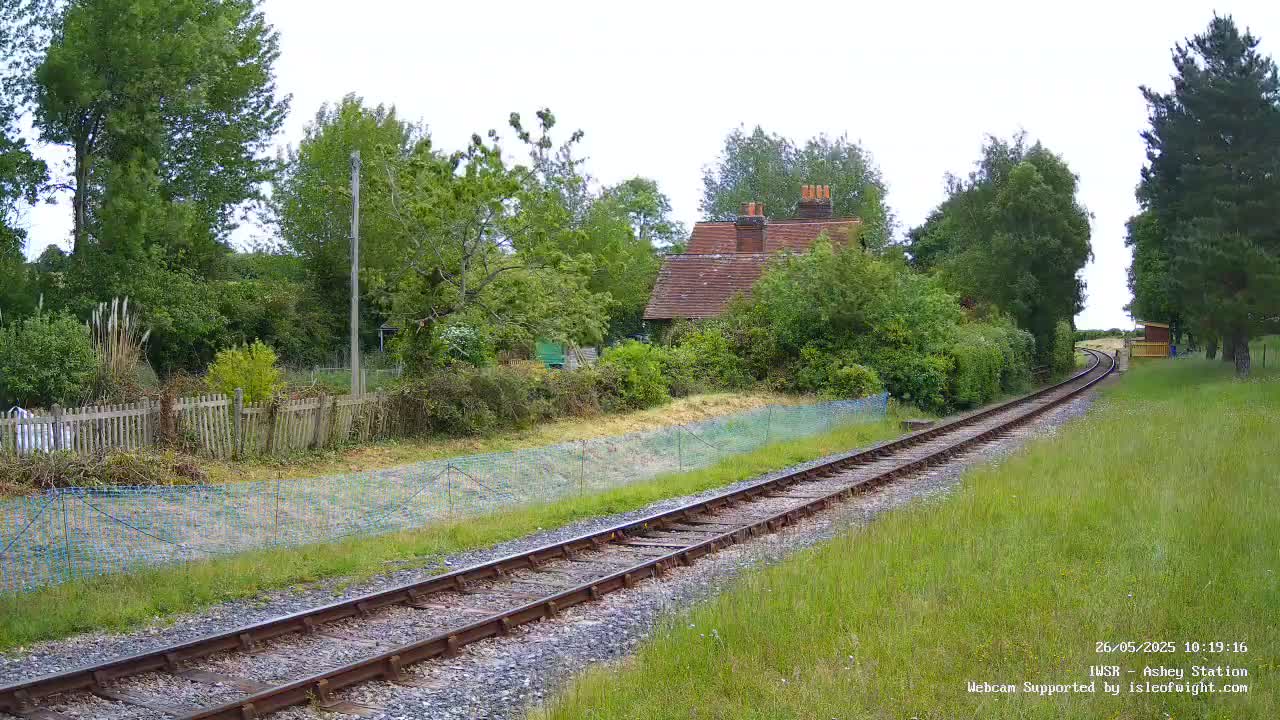 A railway track winds through a verdant landscape, passing a house partially obscured by trees and overgrown vegetation under a cloudy sky.