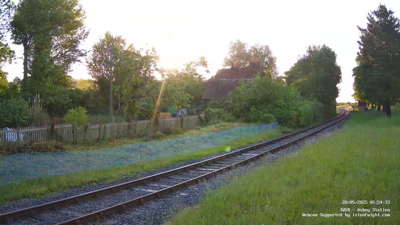 A railway track curves gently through a sunlit grassy field, past a wooden fence and a small house nestled among trees.